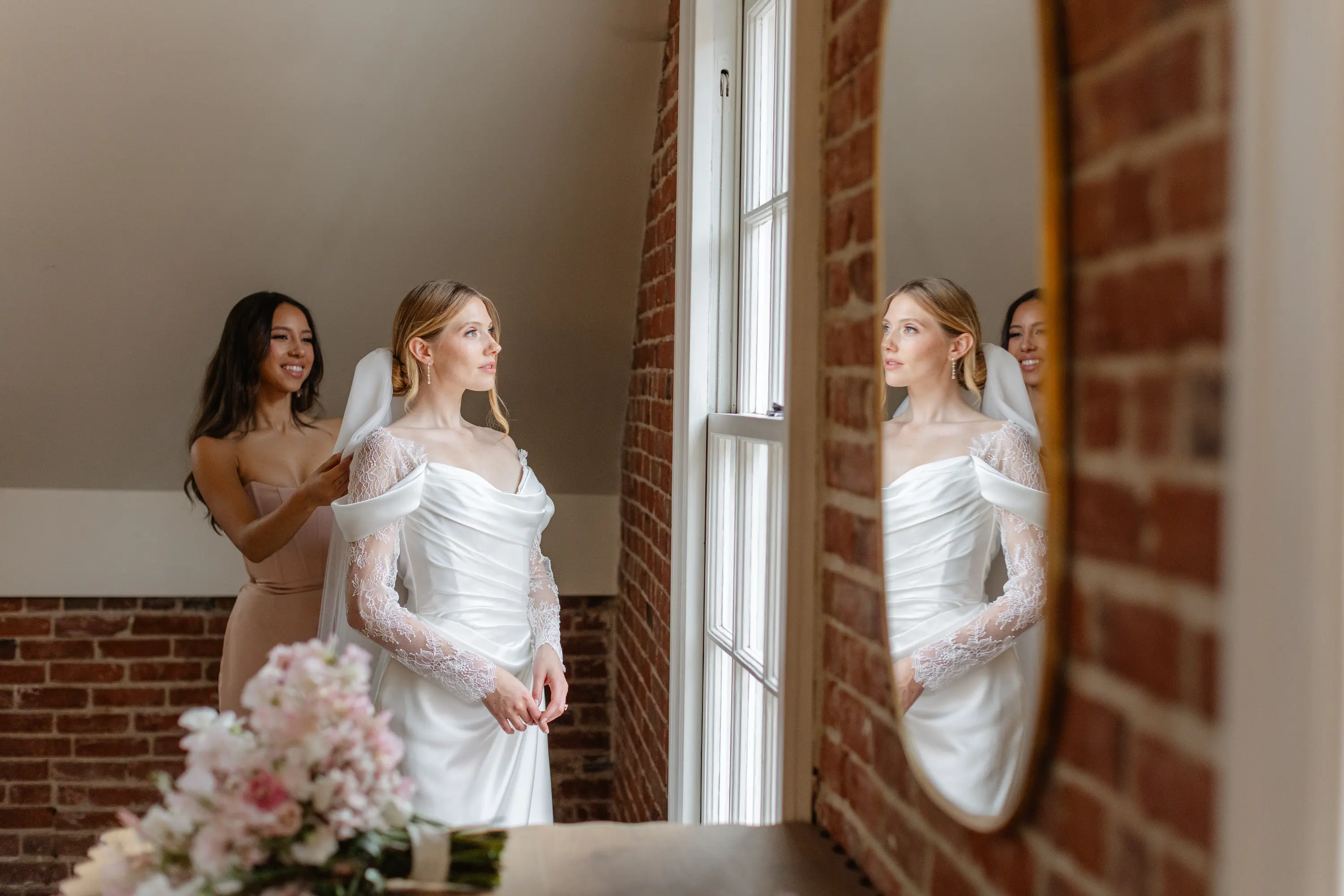 Bride in white lace wedding dress looking out a window while bridesmaid adjusts her veil in a brick-walled room.