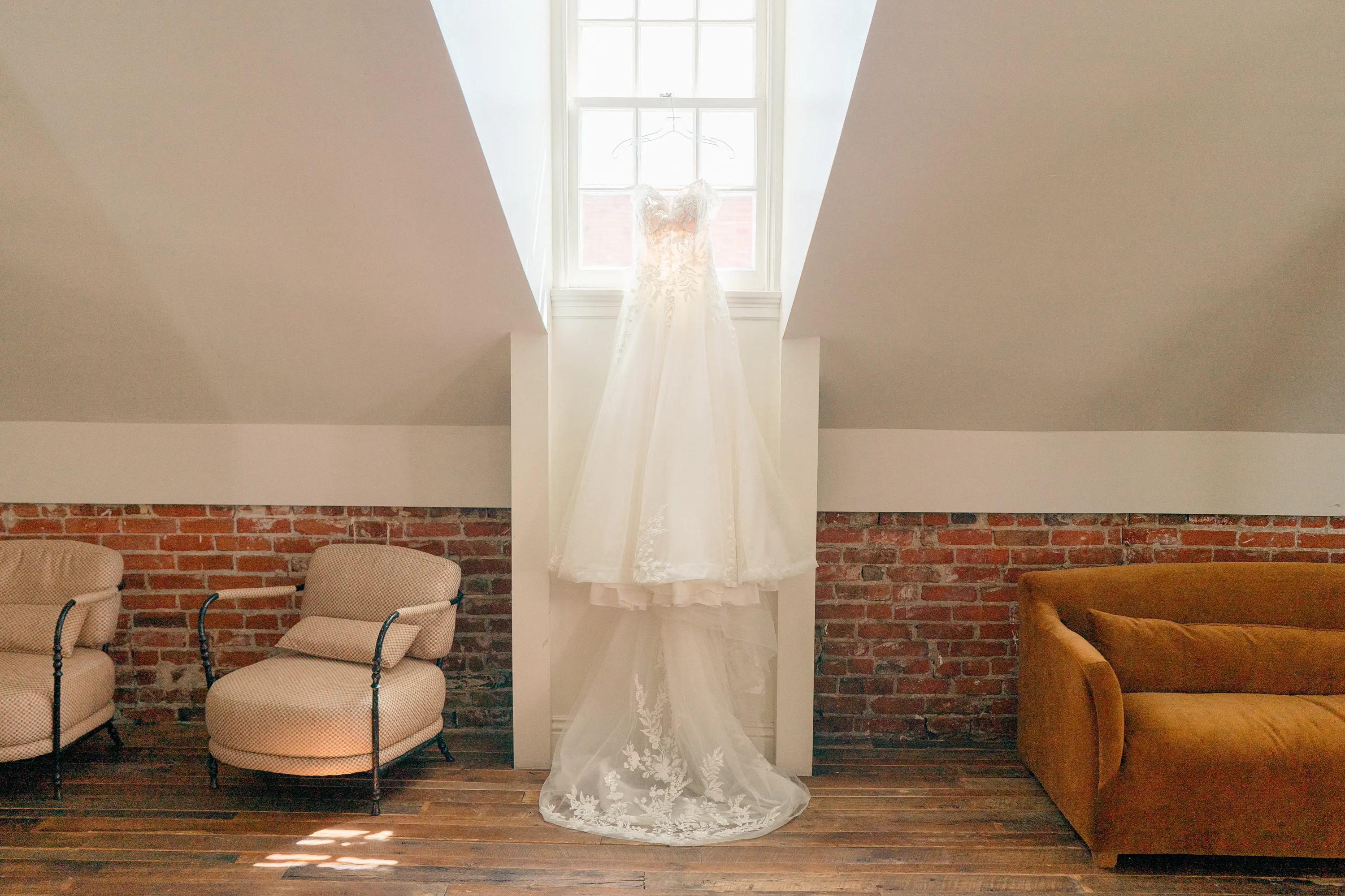White lace wedding dress hanging in a bright room with exposed brick walls and vintage furniture.
