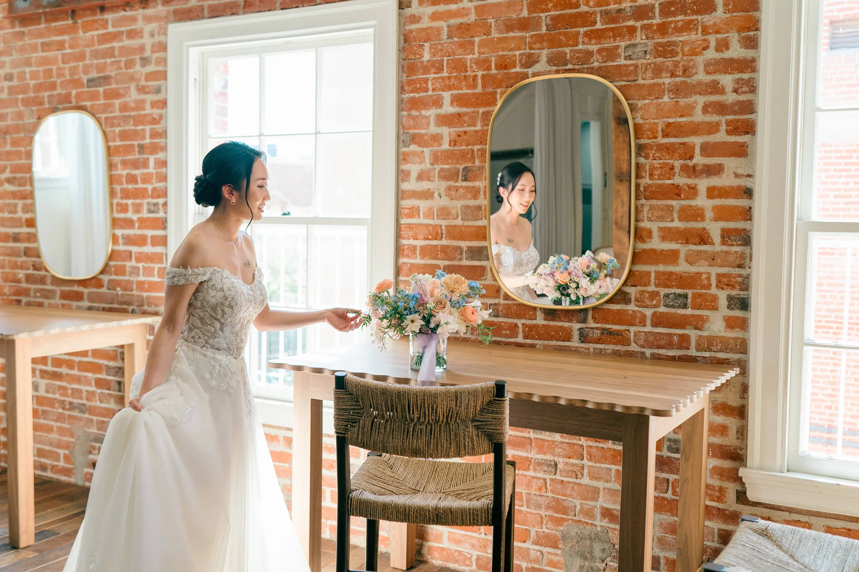 Bride in lace wedding dress standing beside a wooden table with a floral bouquet, smiling at her reflection in a mirror on a brick wall.