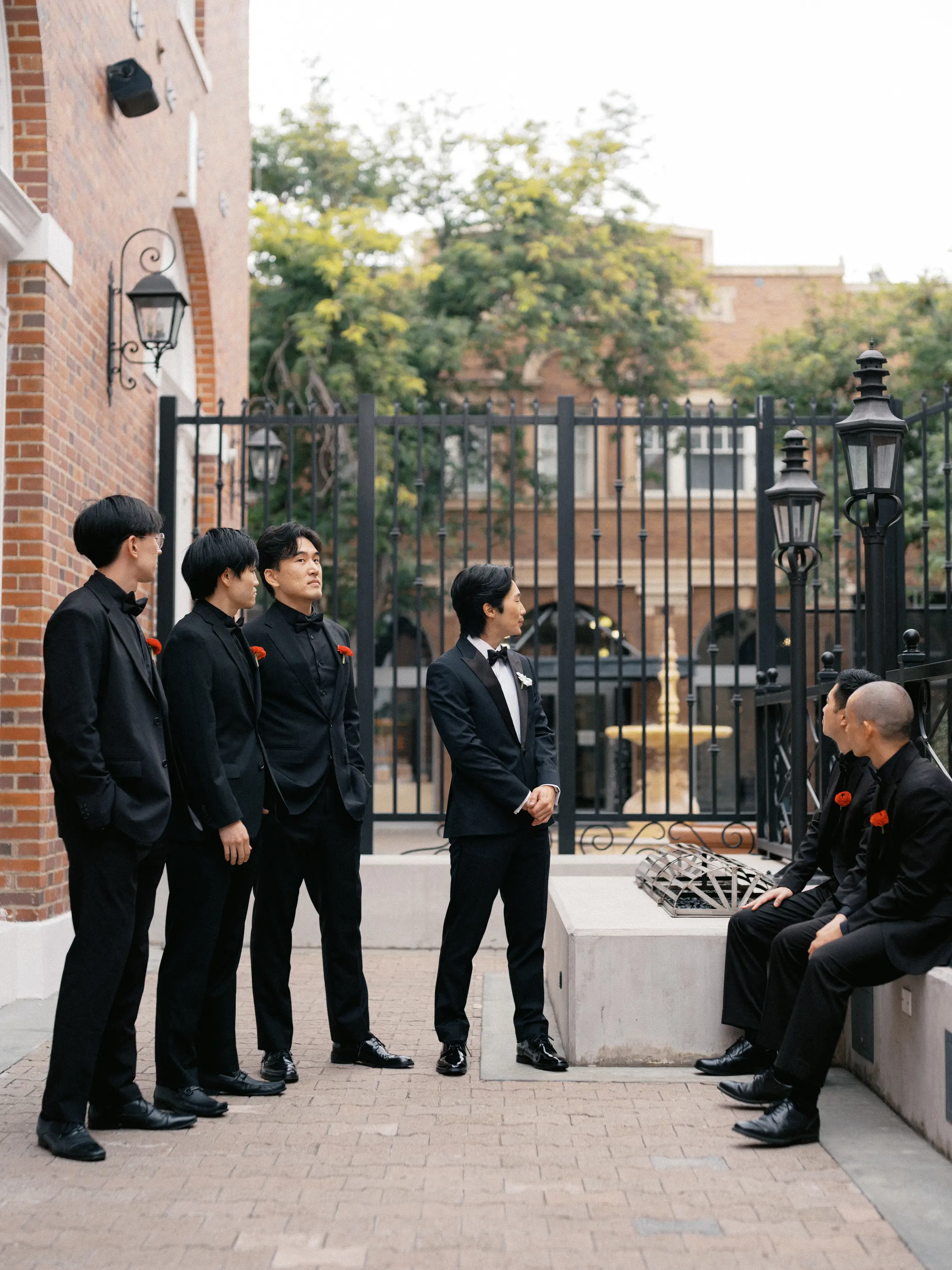 Group of six men dressed in black suits with red boutonnieres, standing and sitting near a wrought iron fence with a brick building and trees in the background.