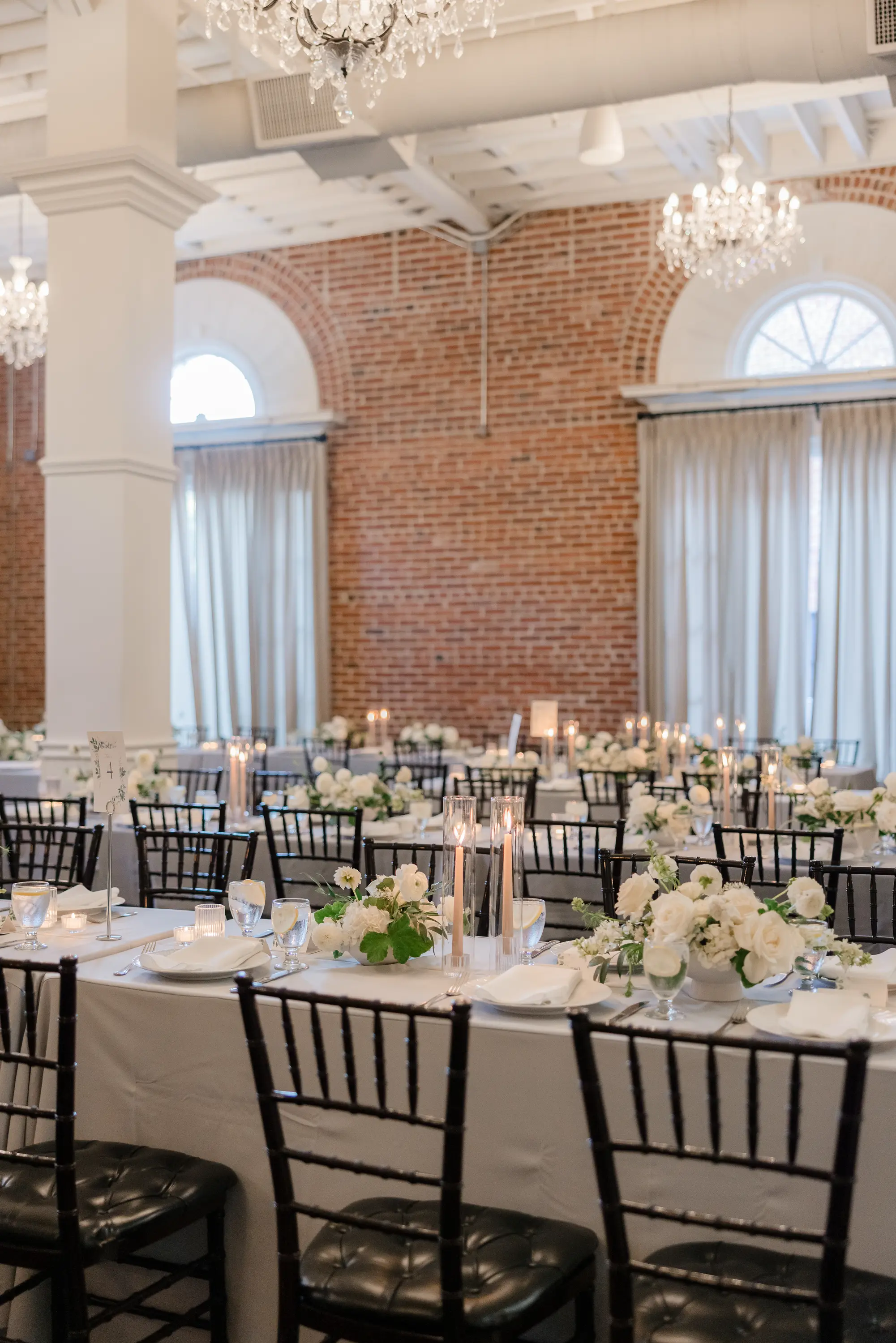 Elegant banquet hall with white floral centerpieces, lit candles, and black chairs arranged around tables with white tablecloths.