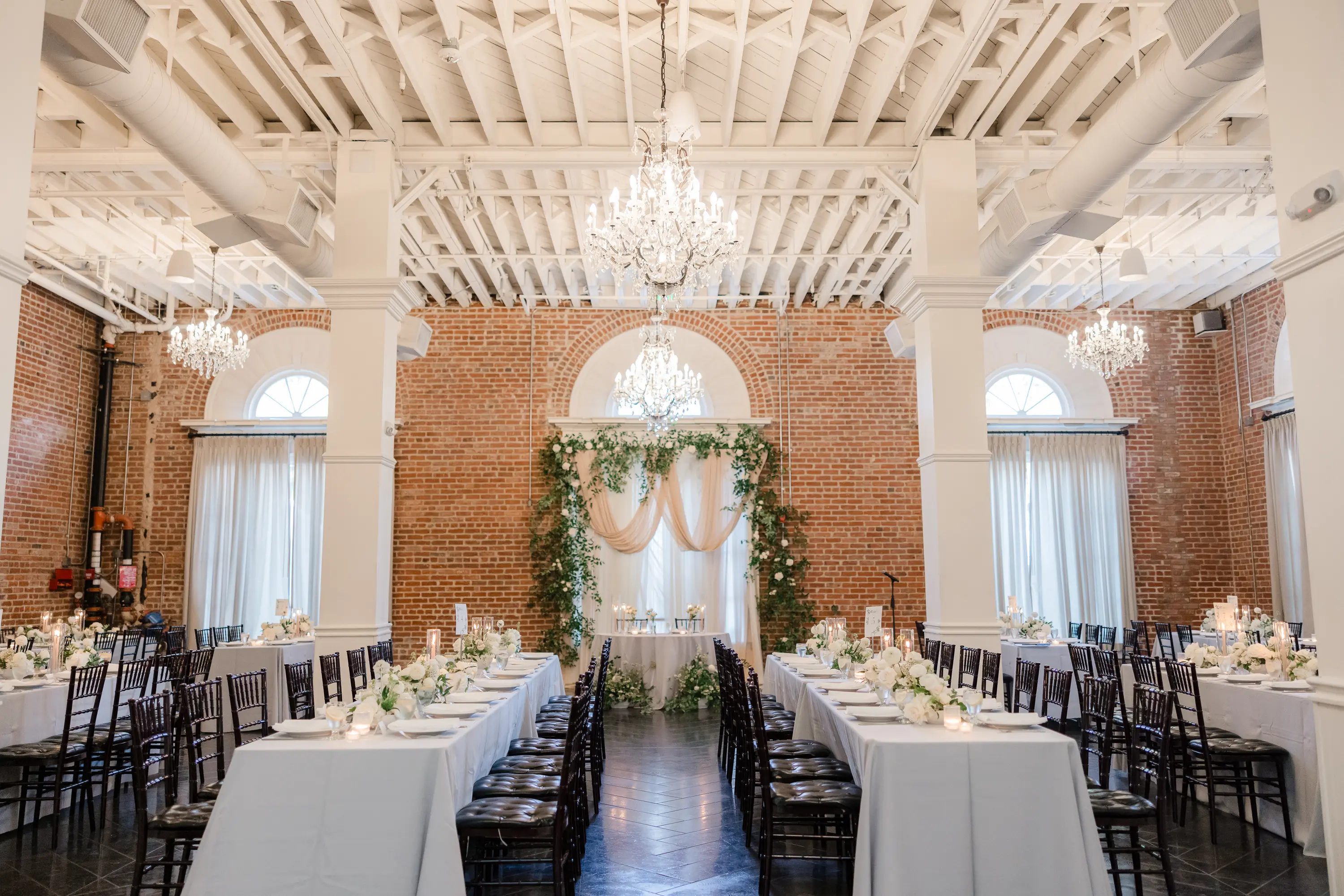 Elegant banquet hall with exposed brick walls, white draped tables, black chairs, and crystal chandeliers.