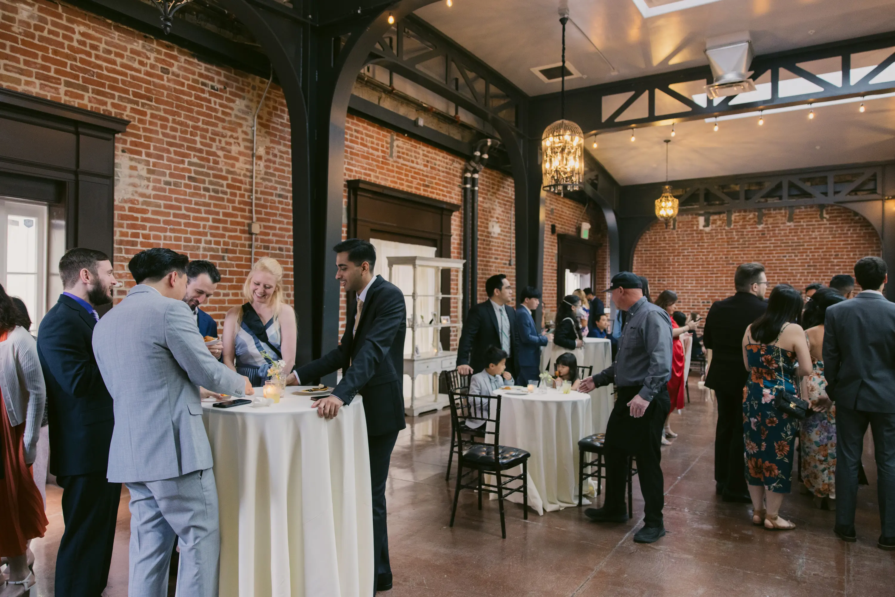 A group of people socializing at a formal indoor event with brick walls, standing and sitting around tables with white tablecloths.