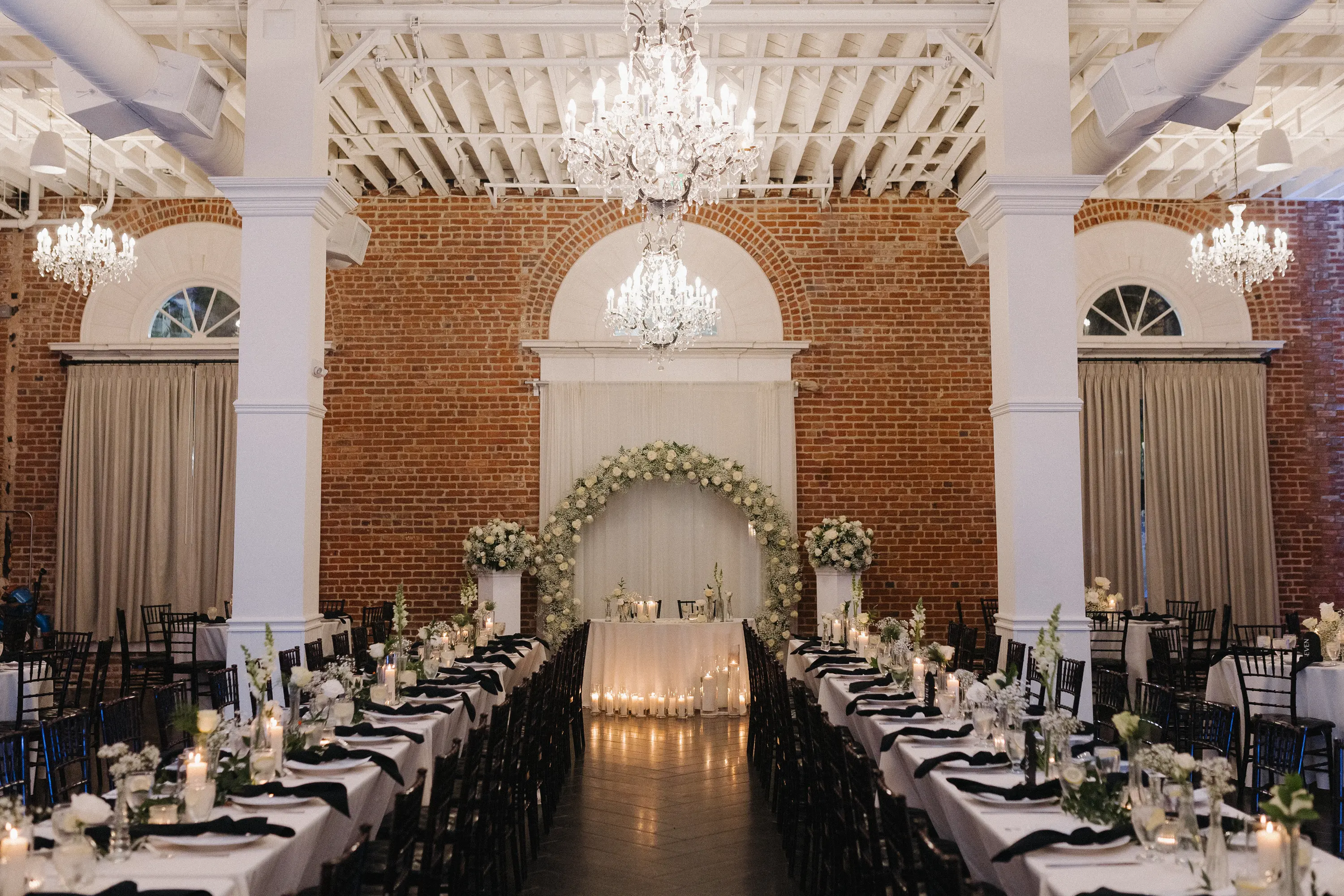 Elegant wedding reception setup with long tables, black chairs, floral centerpieces, candles, and a floral arch against exposed brick walls and chandeliers.