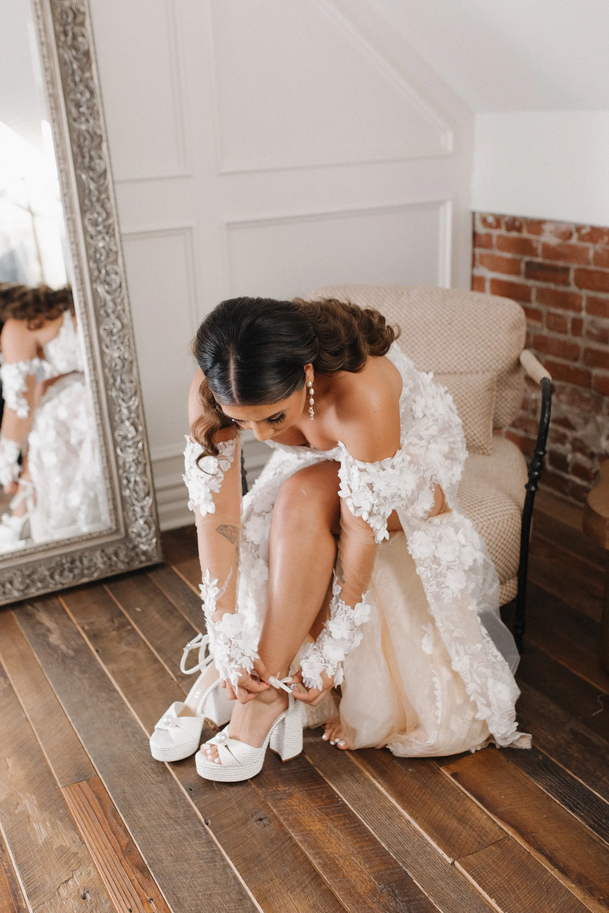 Bride with long, dark hair in a white floral lace wedding gown sitting on a chair and fastening her white platform high heels.