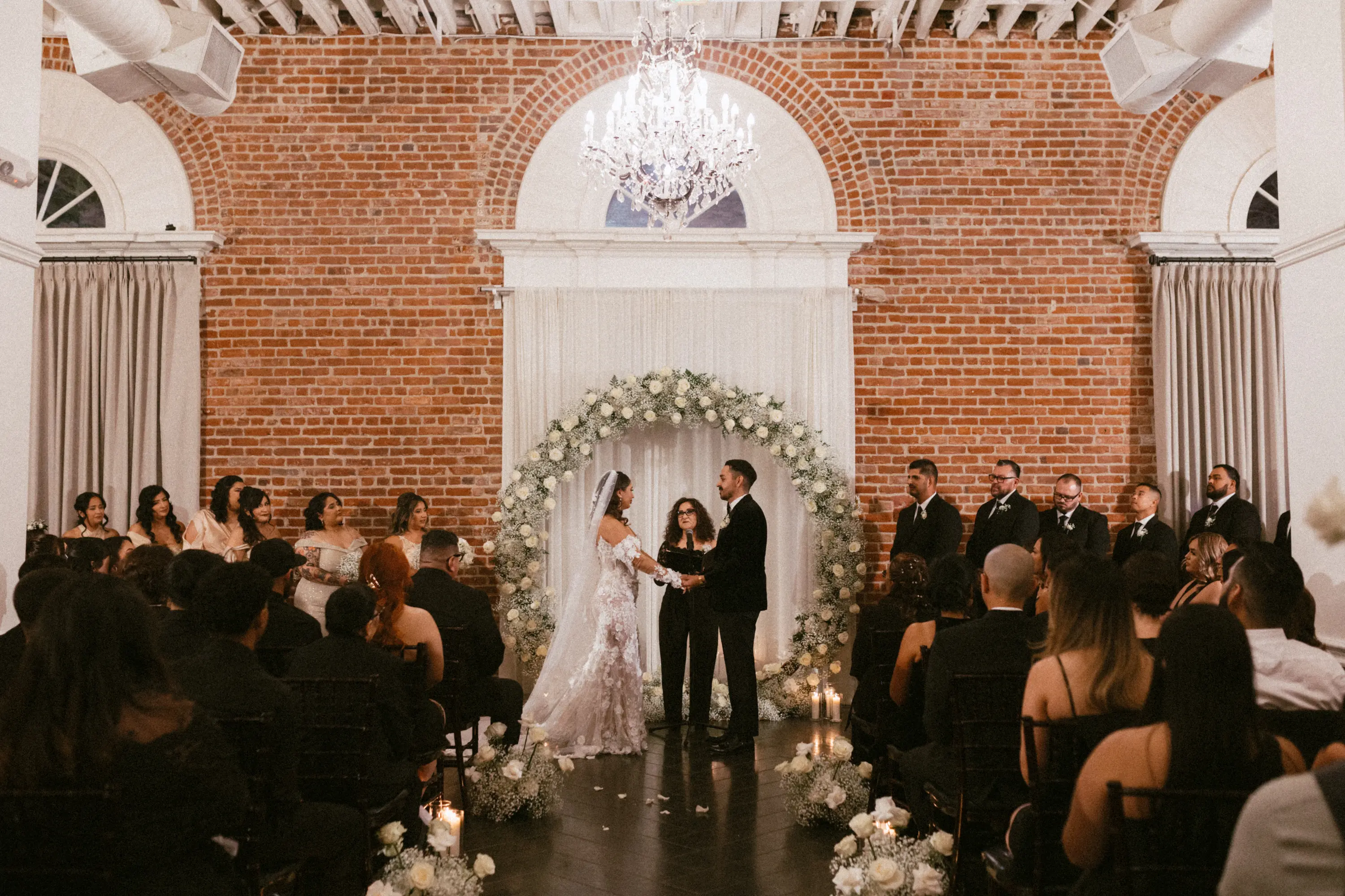 Bride and groom holding hands under a large floral wreath during an indoor wedding ceremony in a room with exposed brick walls and chandelier.