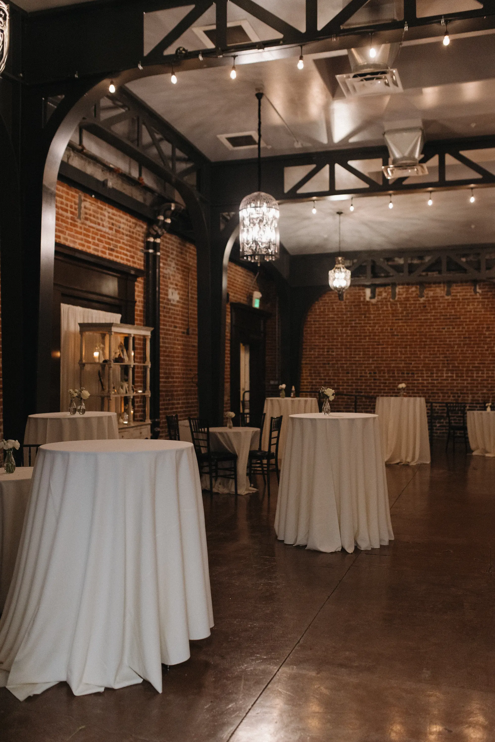 Empty event space with round cocktail tables draped in white cloths, small floral centerpieces, exposed brick walls, and industrial ceiling beams with hanging lights.