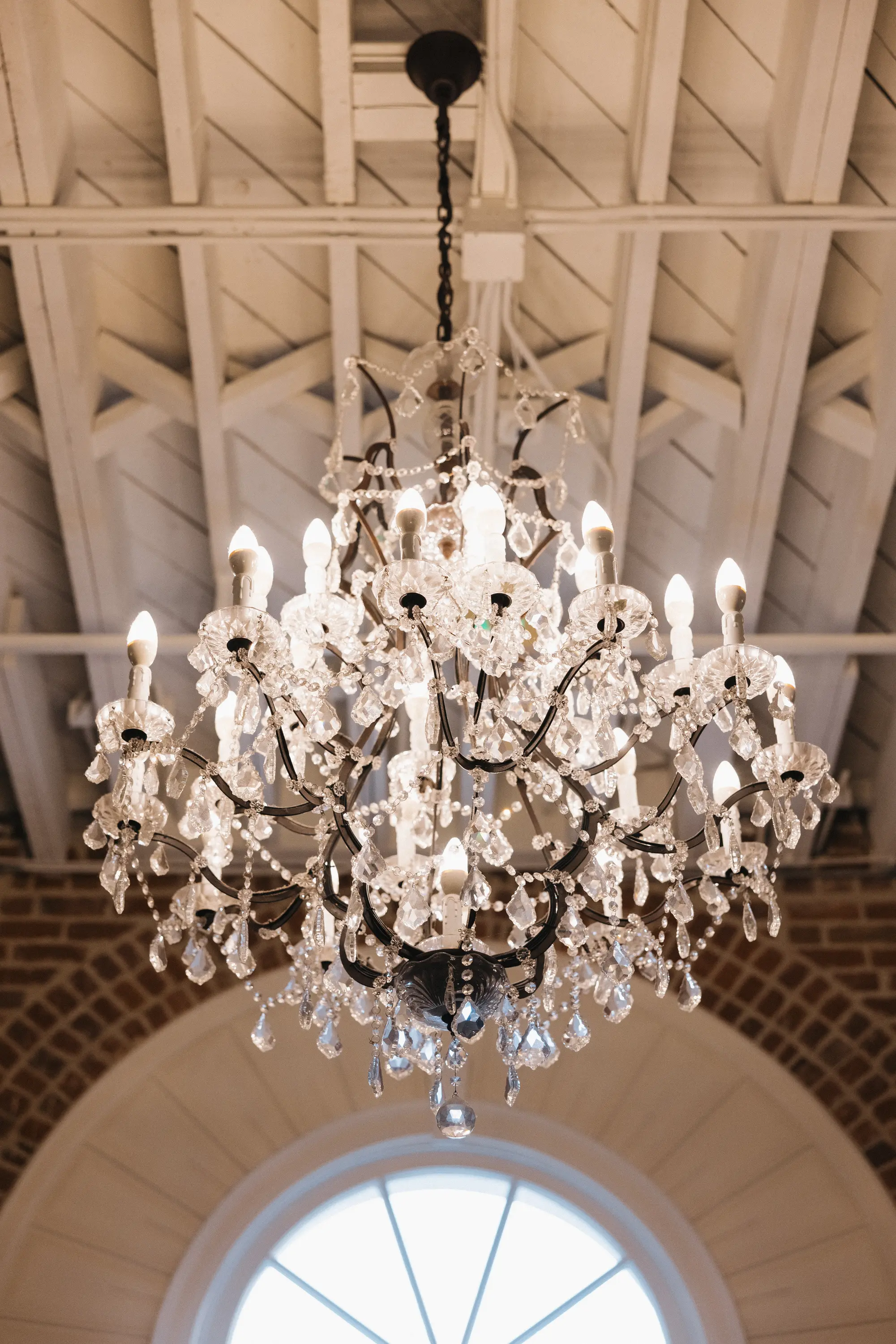 Elegant crystal chandelier with lit bulbs hanging from a white beamed ceiling above an arched window.