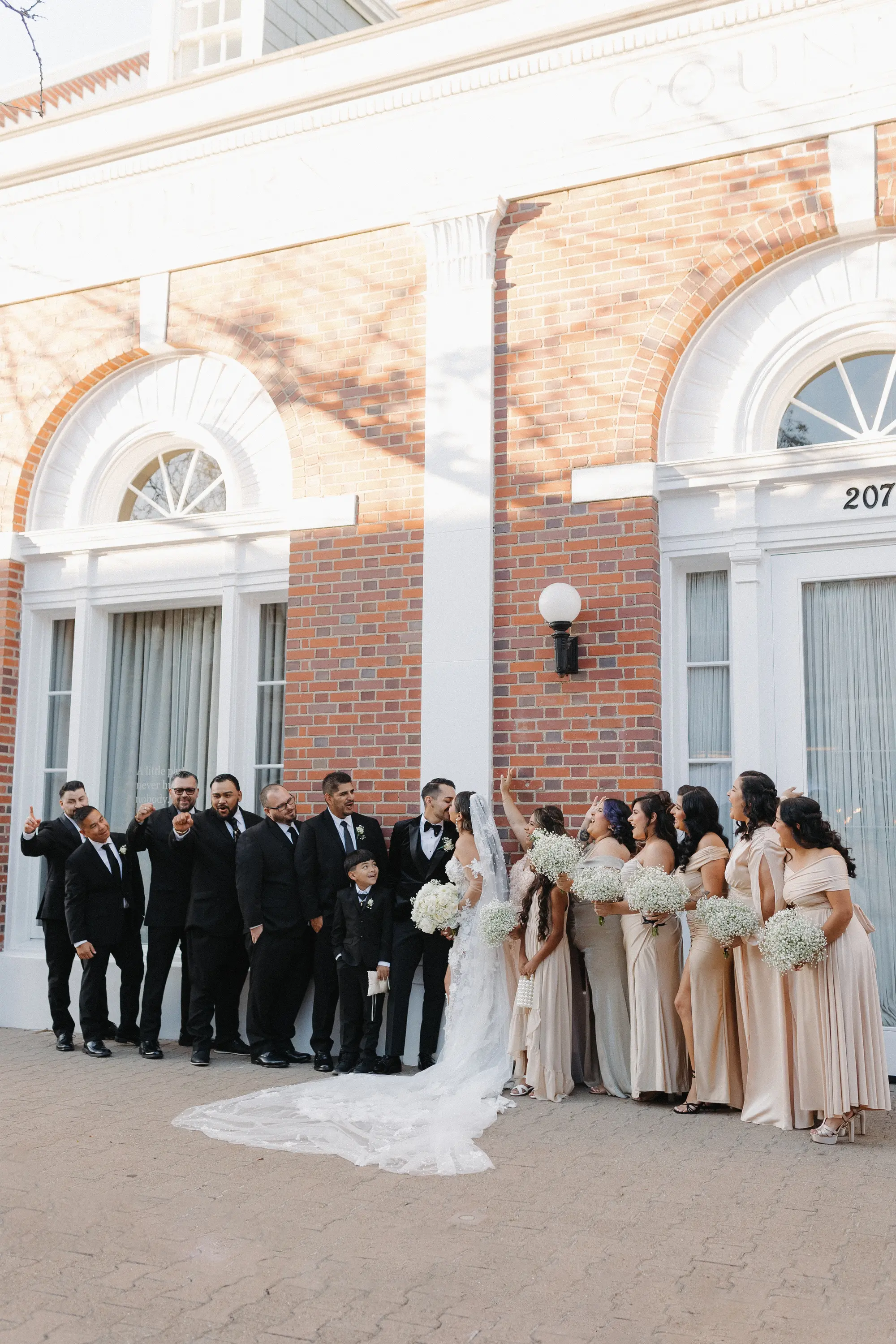 Bride and groom kissing surrounded by bridesmaids in beige dresses and groomsmen in black suits outside a brick building.