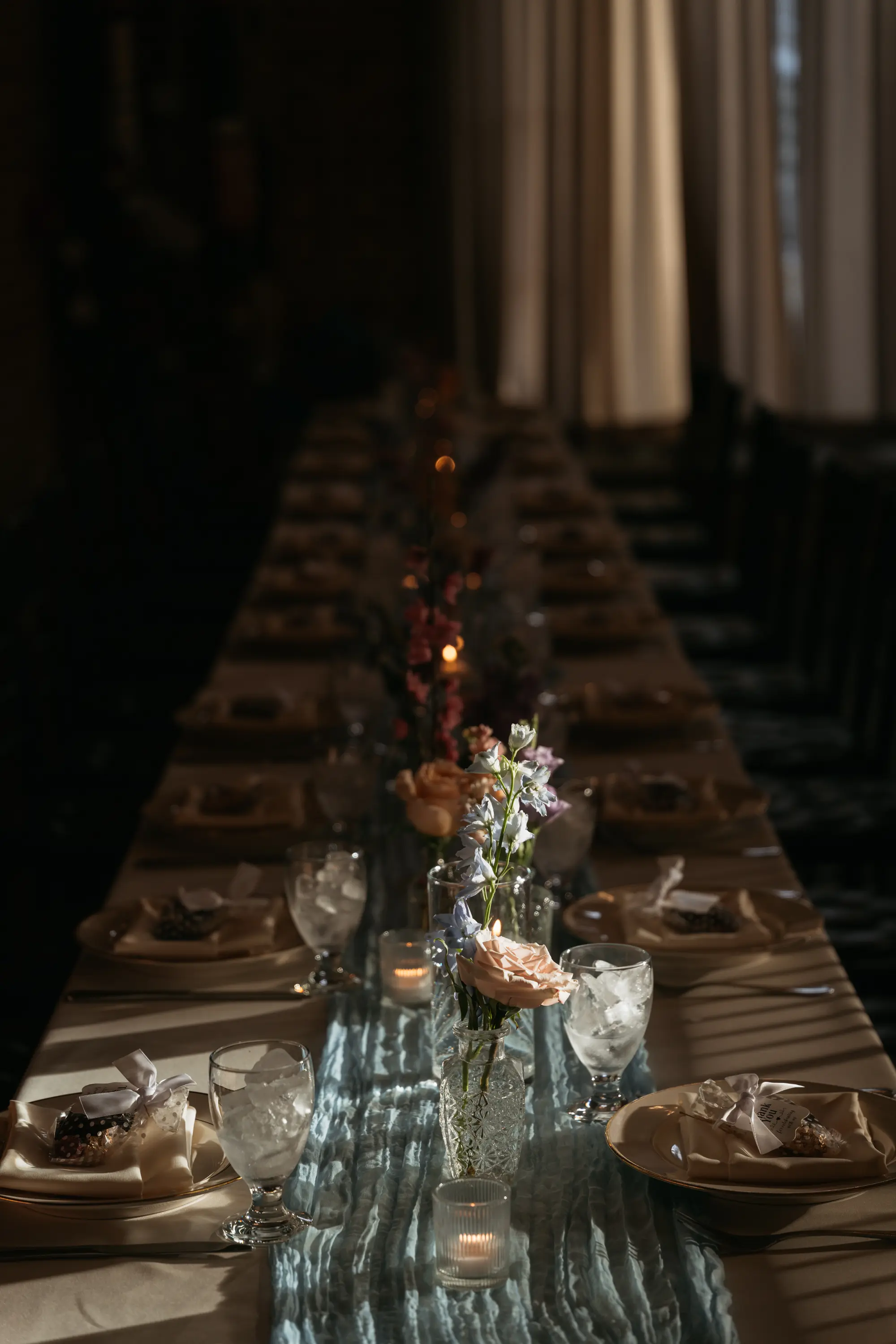 Long dining table elegantly set with plates, ice-filled glasses, candles, and floral centerpieces in soft natural light.