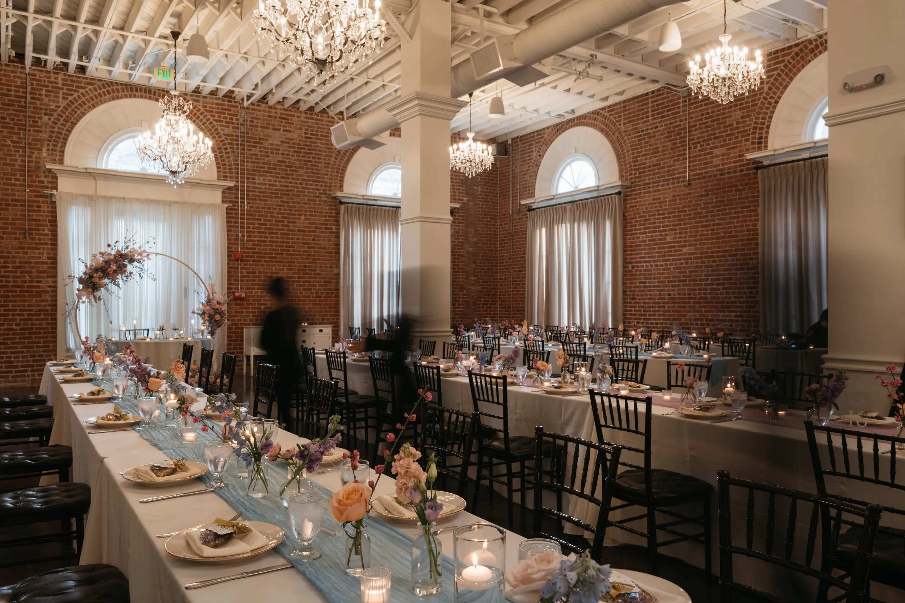 Elegant reception room with brick walls, chandeliers, and long tables set with white tablecloths, flowers, candles, and black chairs.