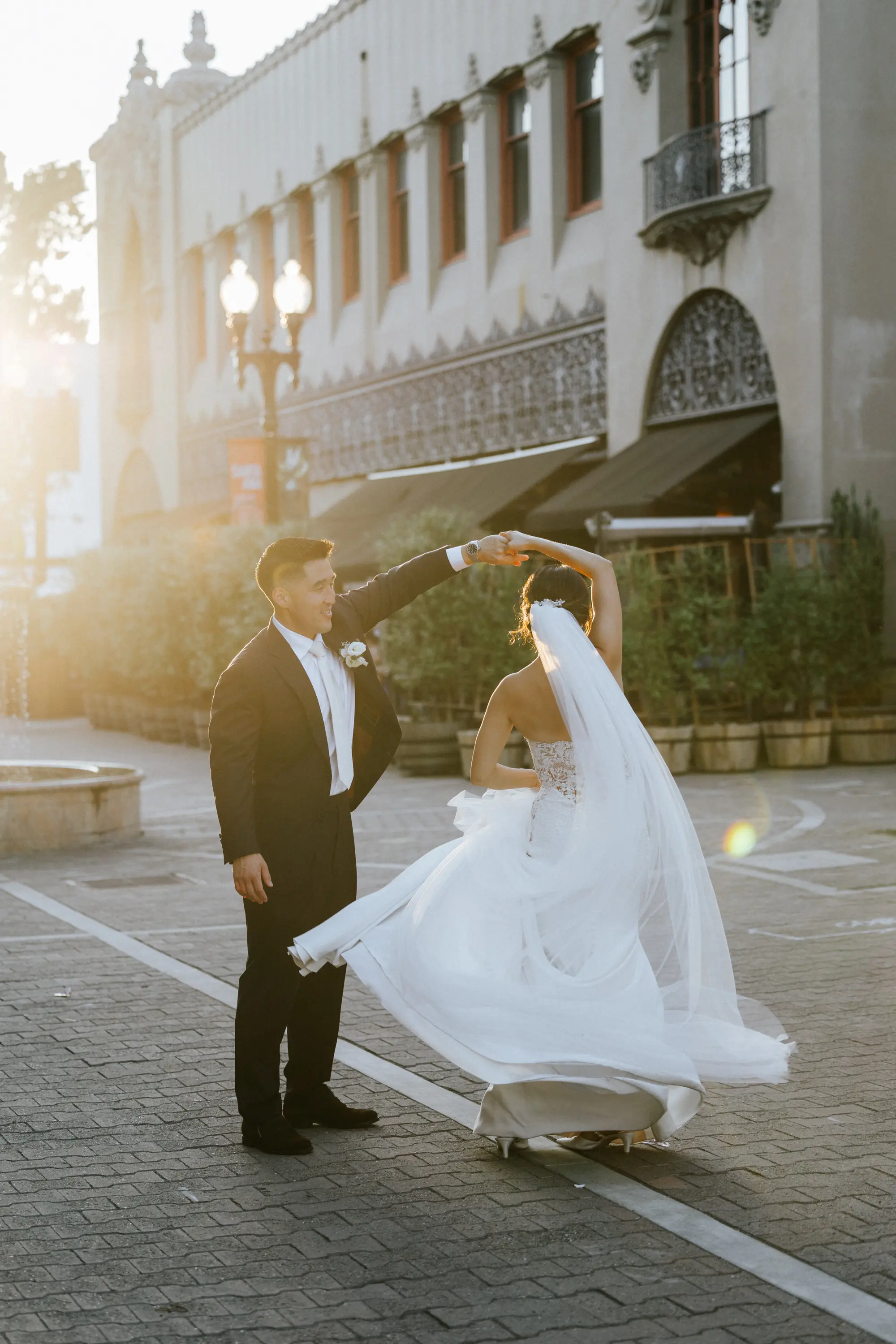 Groom twirling bride in white wedding dress and veil on a sunlit cobblestone street in front of a historic building.
