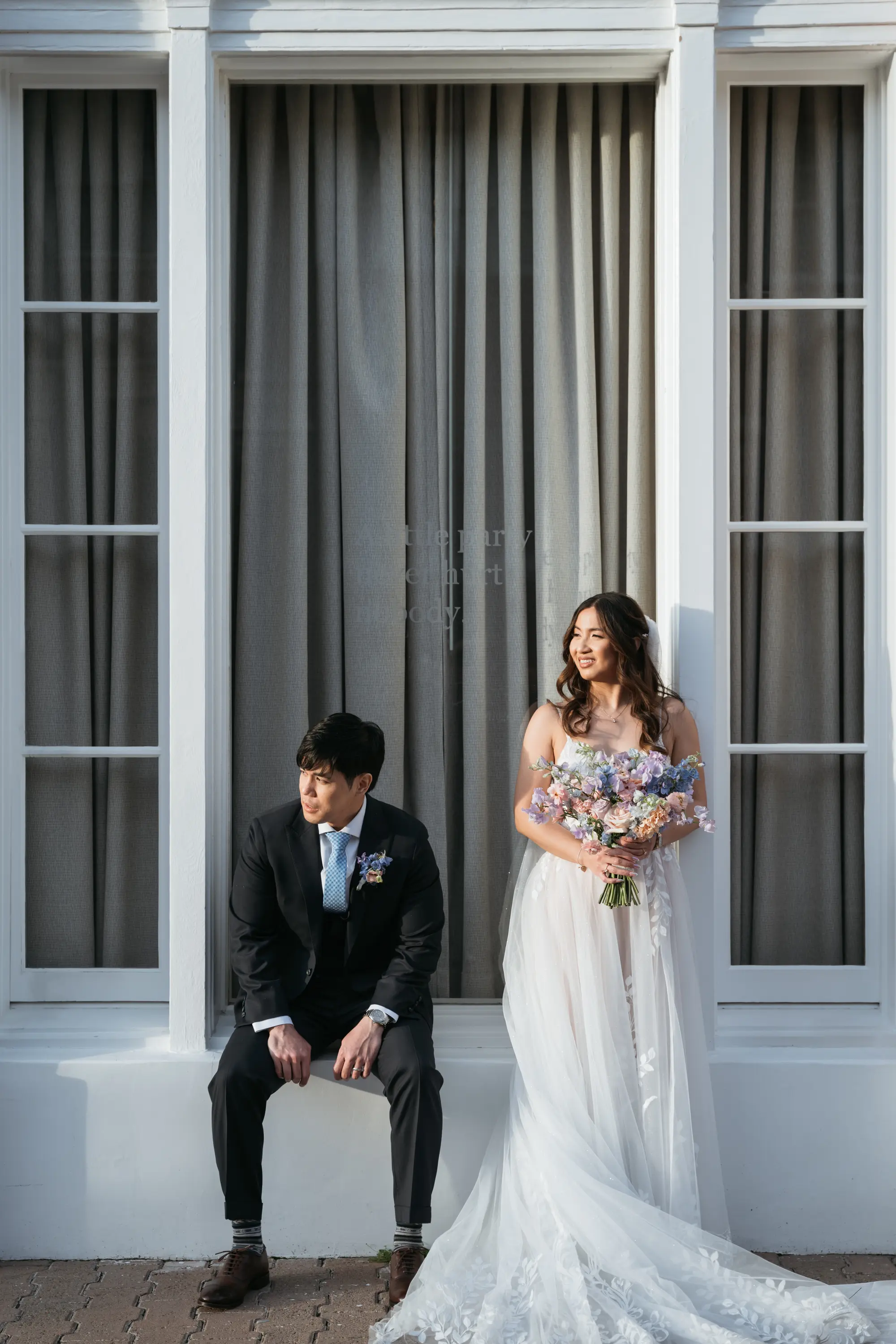 Bride in white wedding dress holding a colorful flower bouquet standing next to groom in black suit sitting on a ledge in front of a large window with gray curtains.