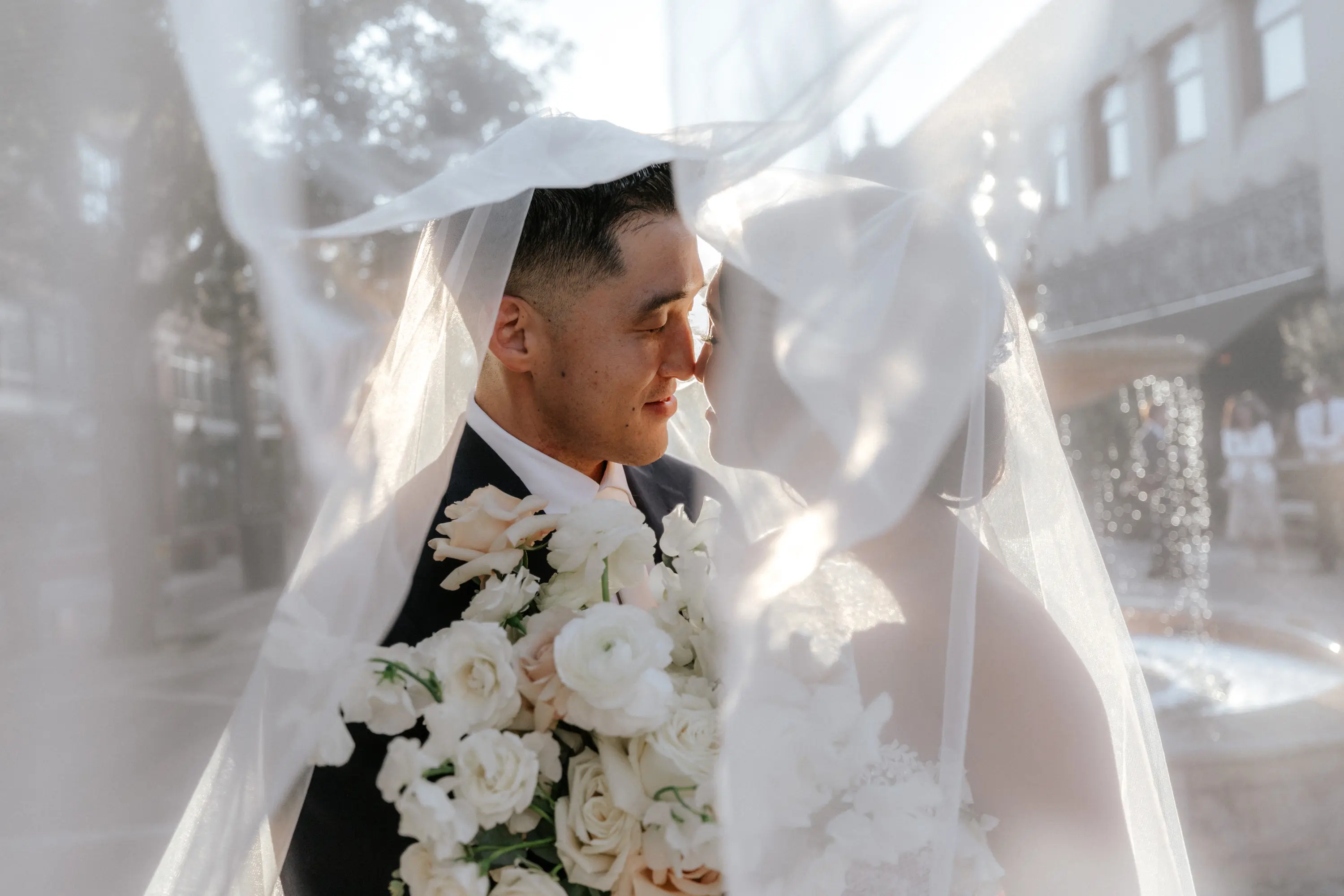 Bride and groom touching noses tenderly under a flowing white veil with a bouquet of white and pale pink flowers.