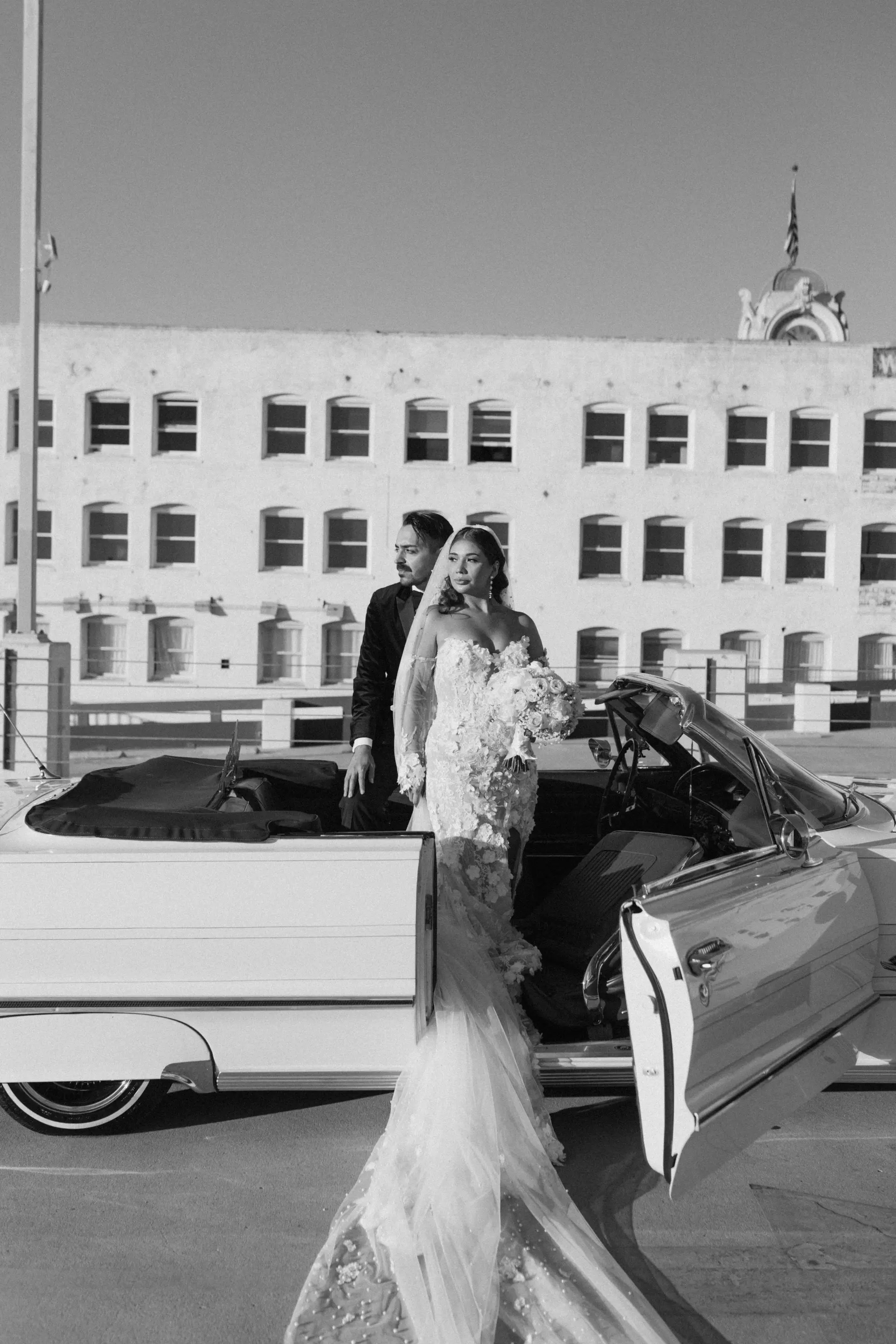 Bride in a long floral lace gown holding a bouquet standing beside an open door of a classic convertible car with a groom in a tuxedo sitting inside, in front of a building with multiple windows.