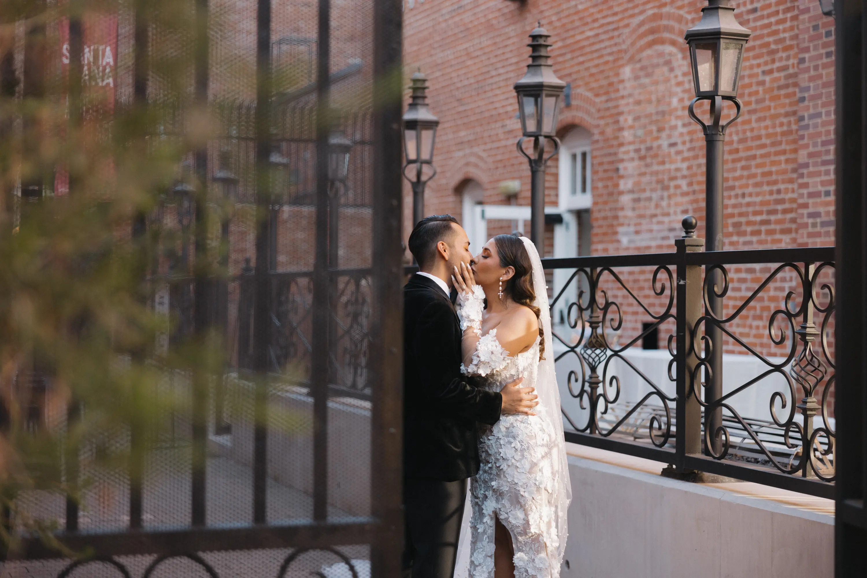Bride and groom dressed in wedding attire share a kiss near decorative black iron fence and brick wall.