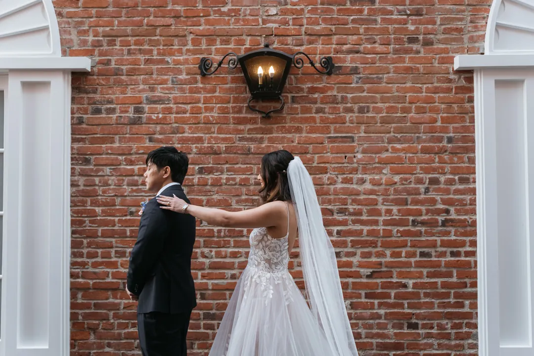 Bride approaching the groom from behind beside a brick wall