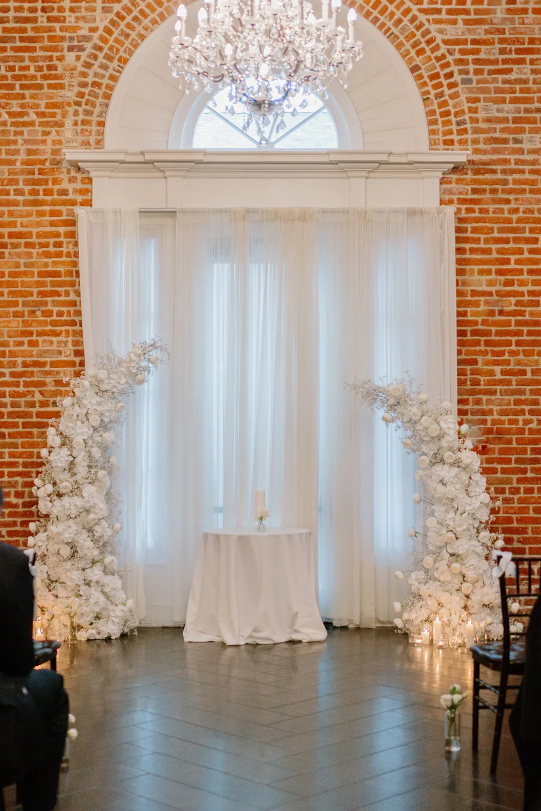 Floral ceremony arrangements beneath a chandelier and arched window