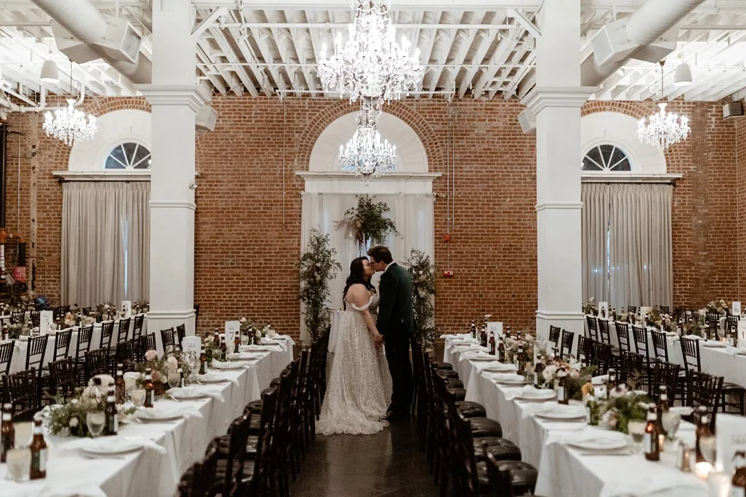 Couple standing between long reception tables beneath chandeliers