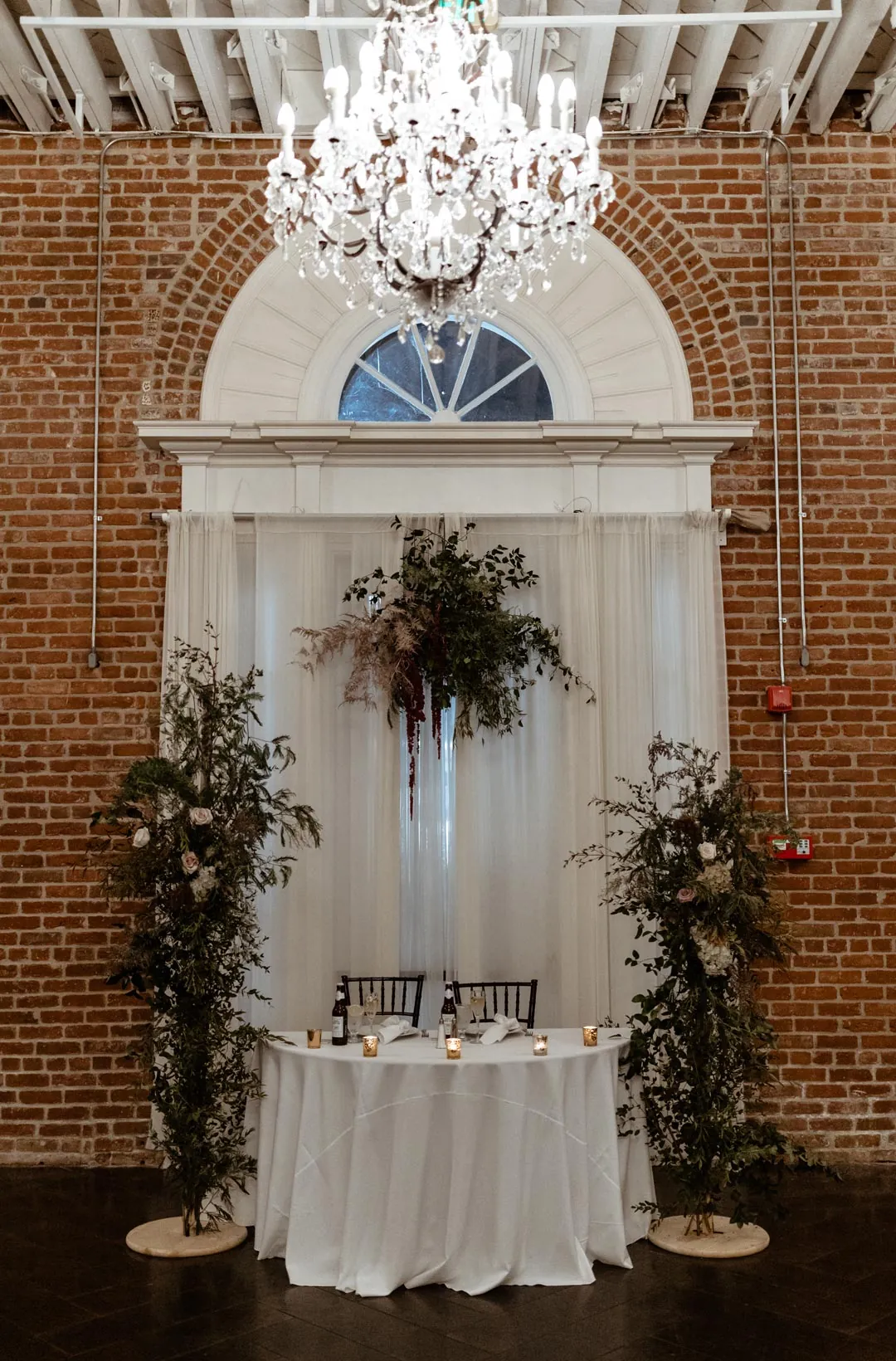 Round sweetheart table beneath a chandelier and arched window