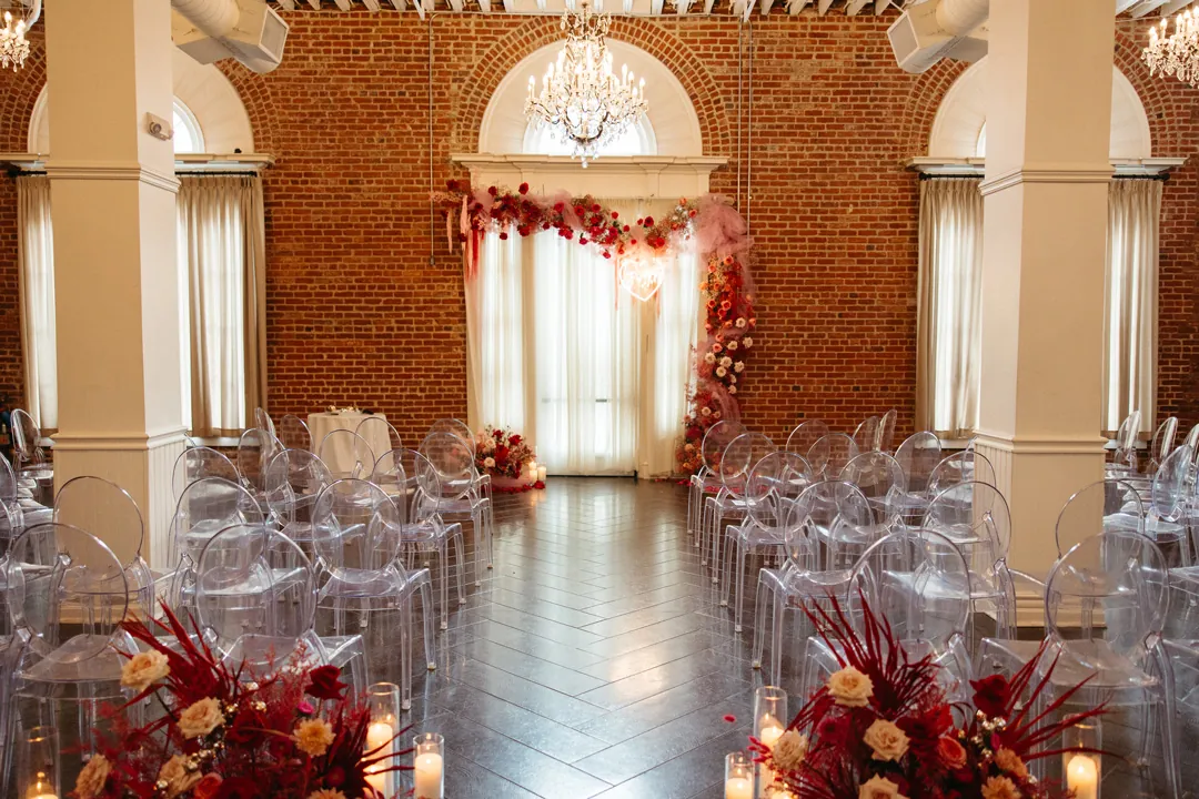Clear ceremony chairs flank a red flower lined aisle.