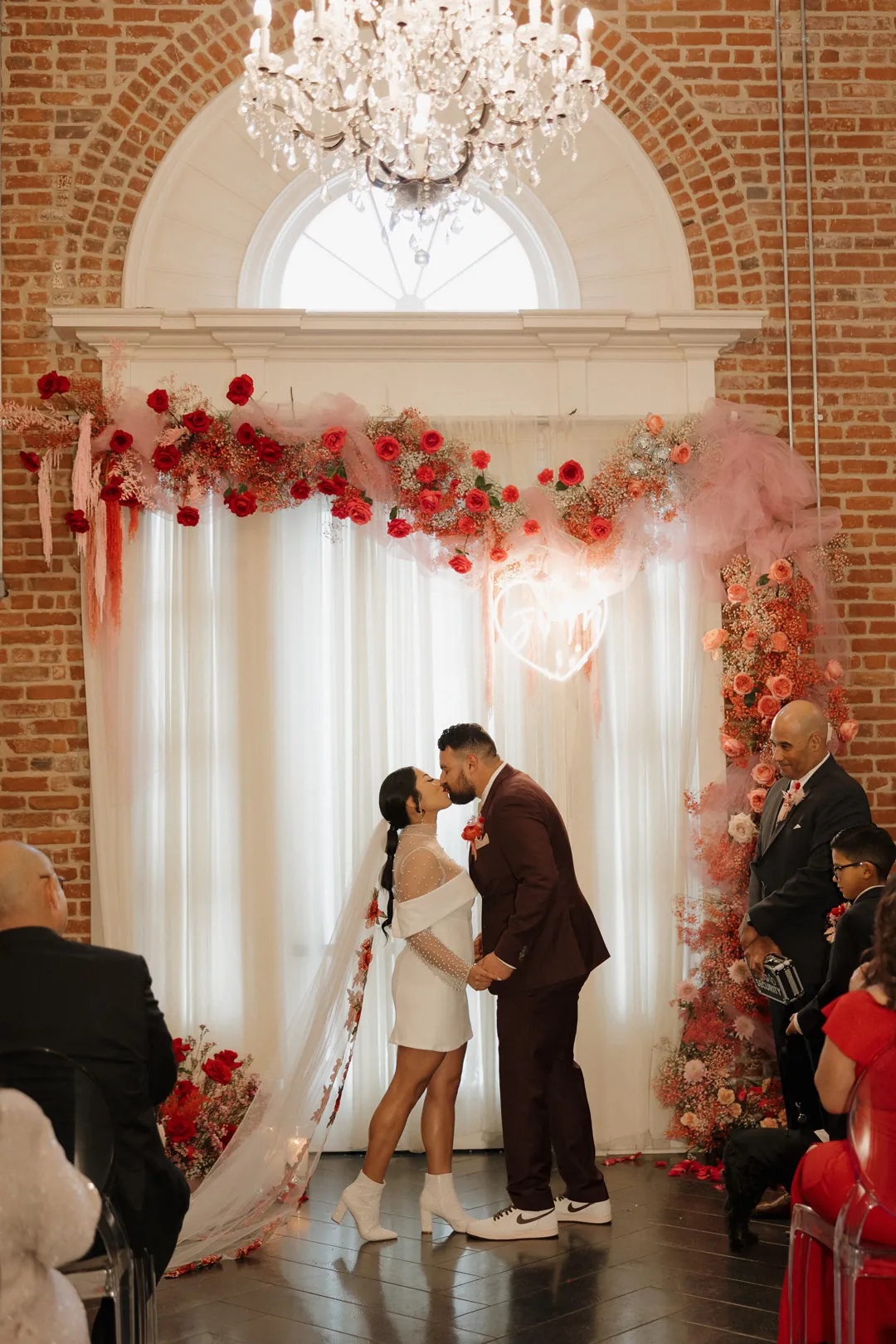 Newlyweds kiss before a red floral ceremony backdrop.