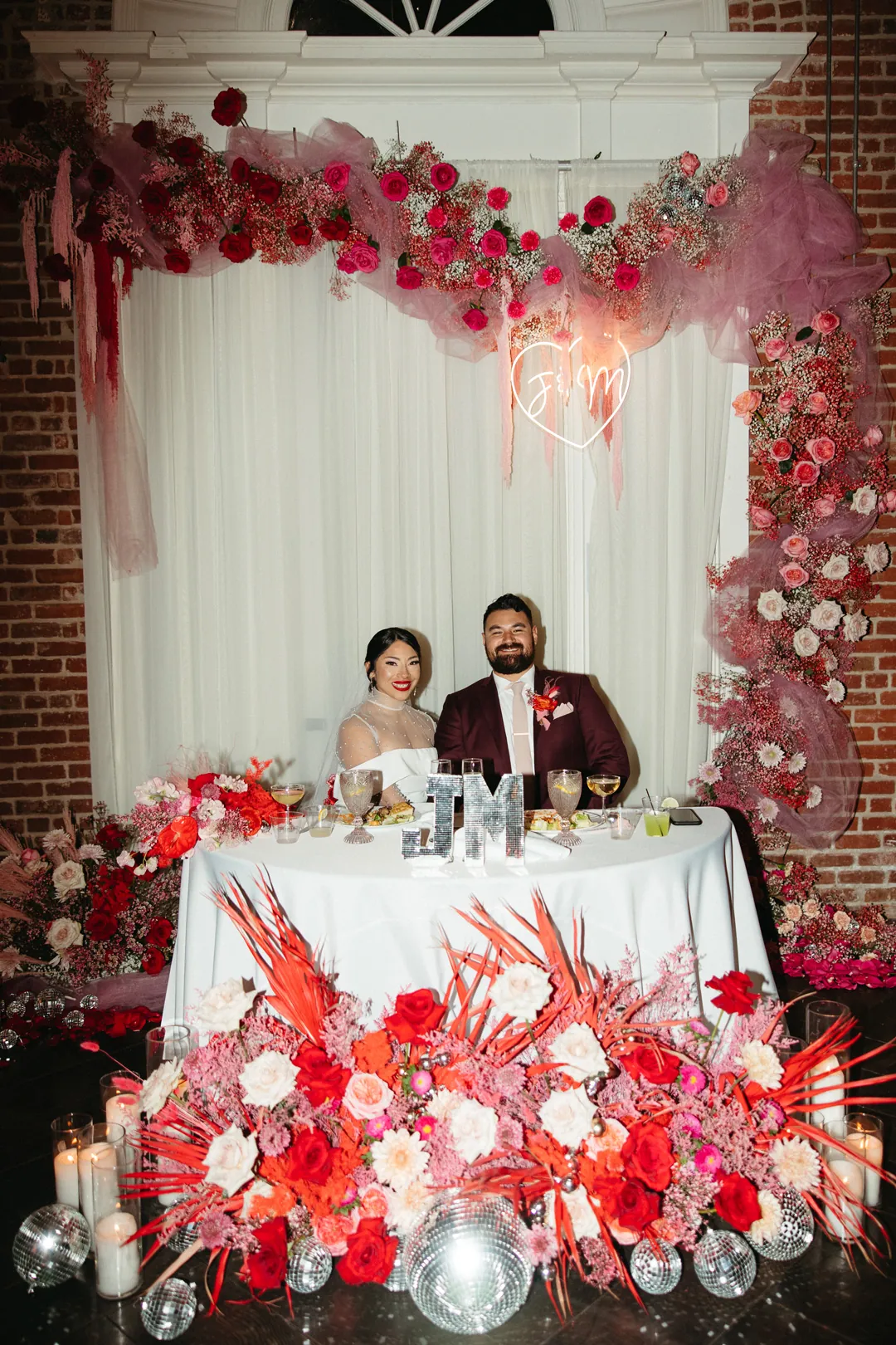 Newlyweds sit beneath a red floral arch.