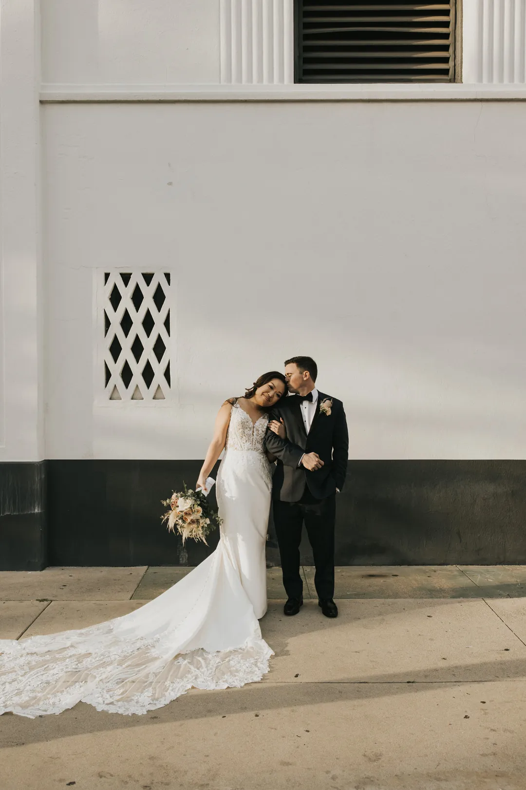 Couple embrace beside a pale exterior wall.