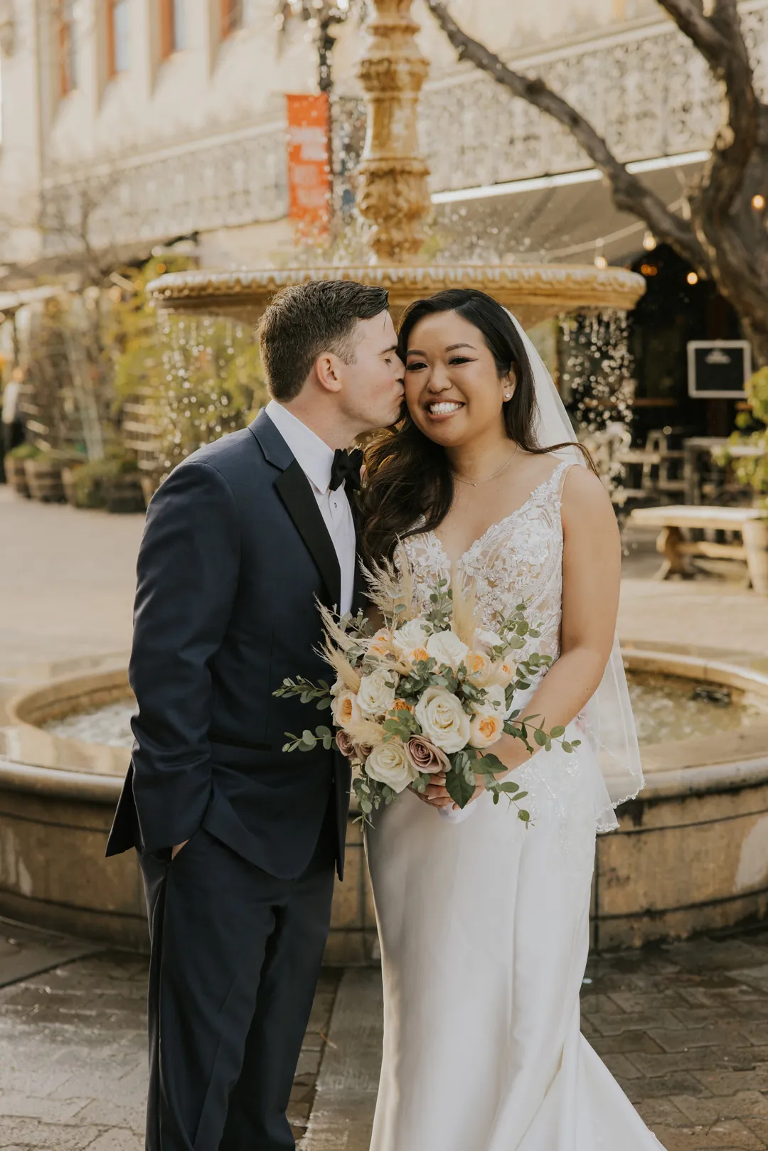 Groom kisses the bride's cheek near an outdoor fountain.