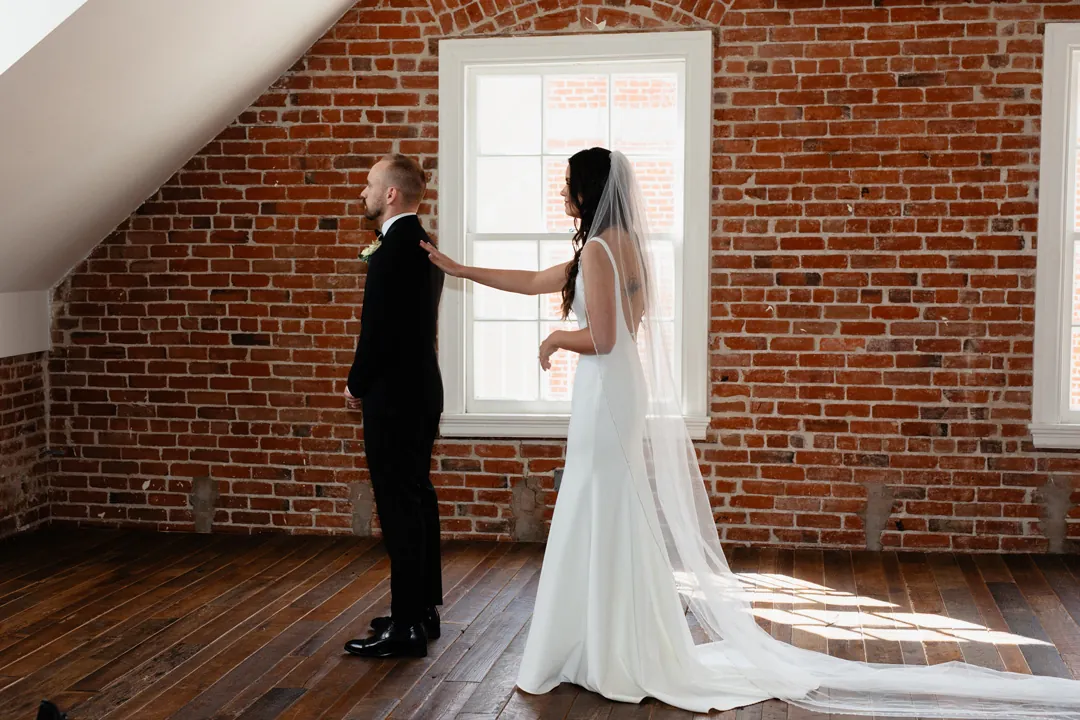 Bride reaching toward the groom beside a window and brick wall