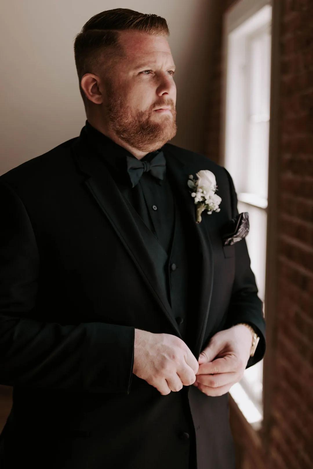 Groom adjusts his black jacket beside a window.