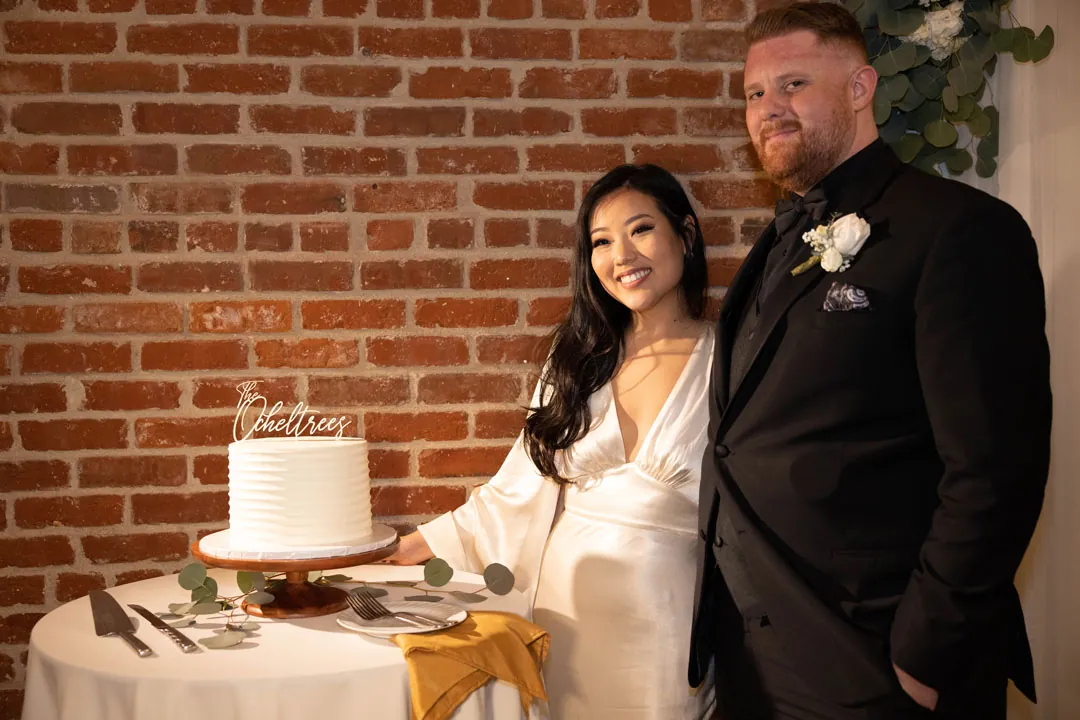 Couple stand beside their wedding cake against exposed brick.