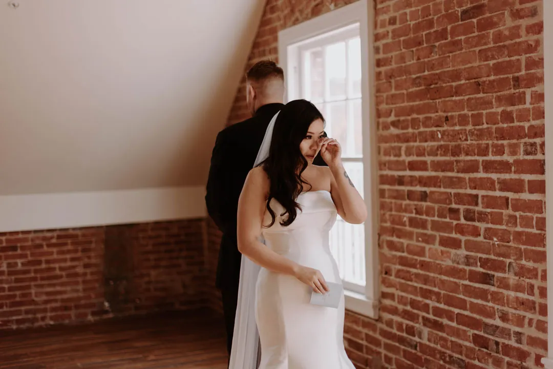 Bride touches her face while standing beside a window.
