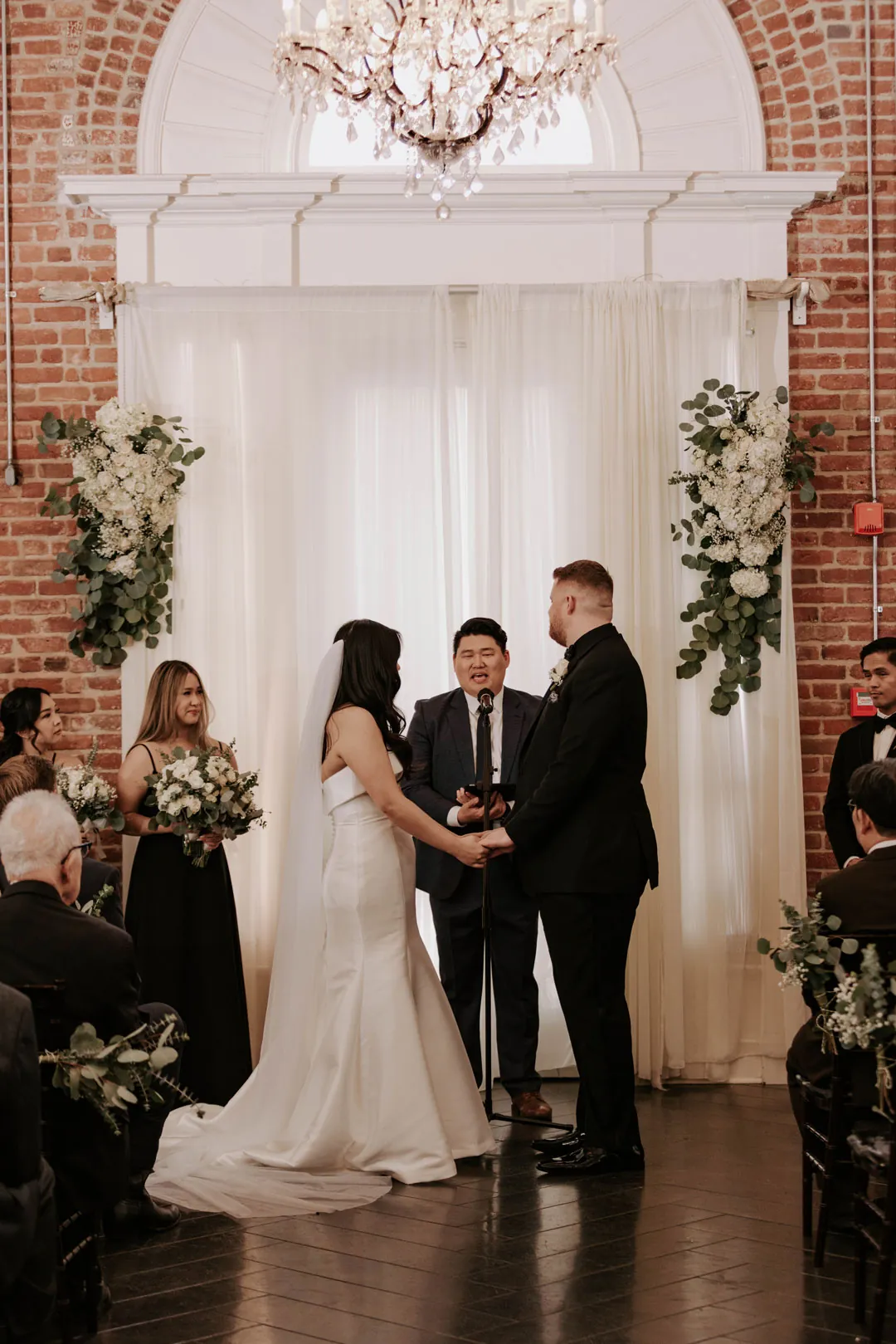 Couple exchange vows before a draped floral backdrop.