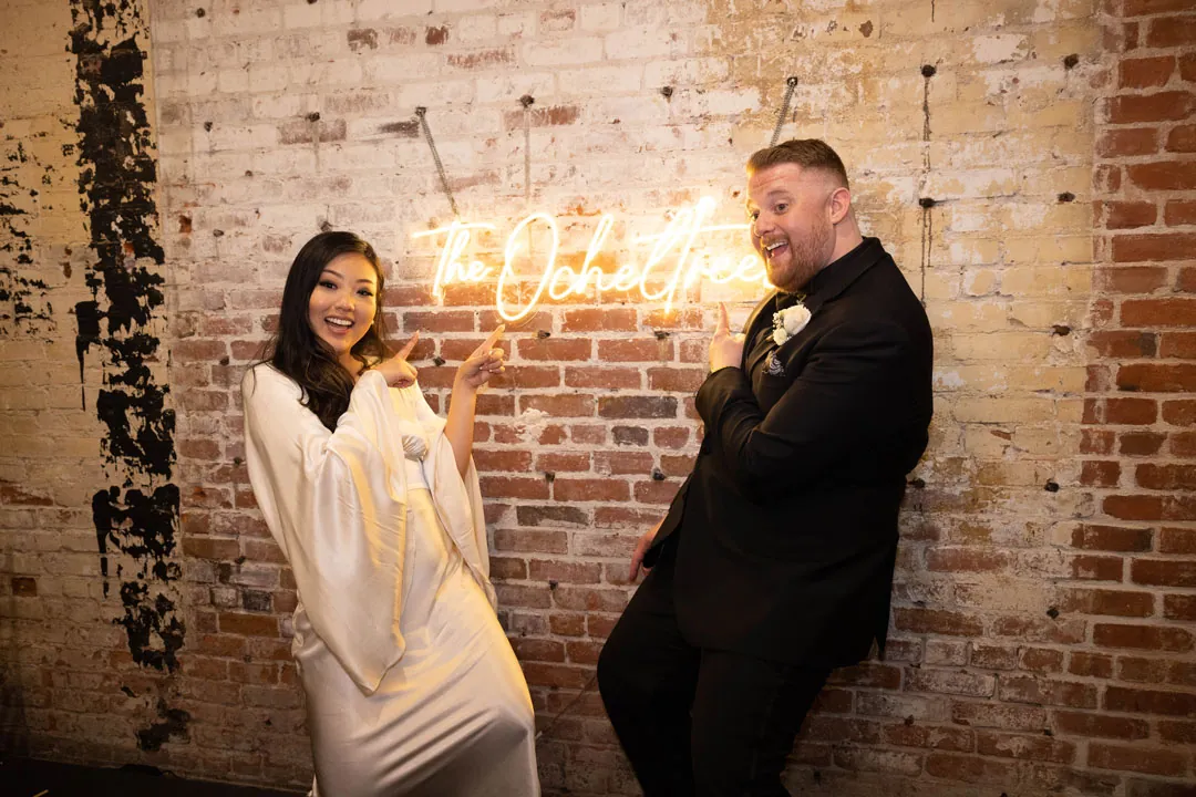 Couple pose against exposed brick beside a neon sign.