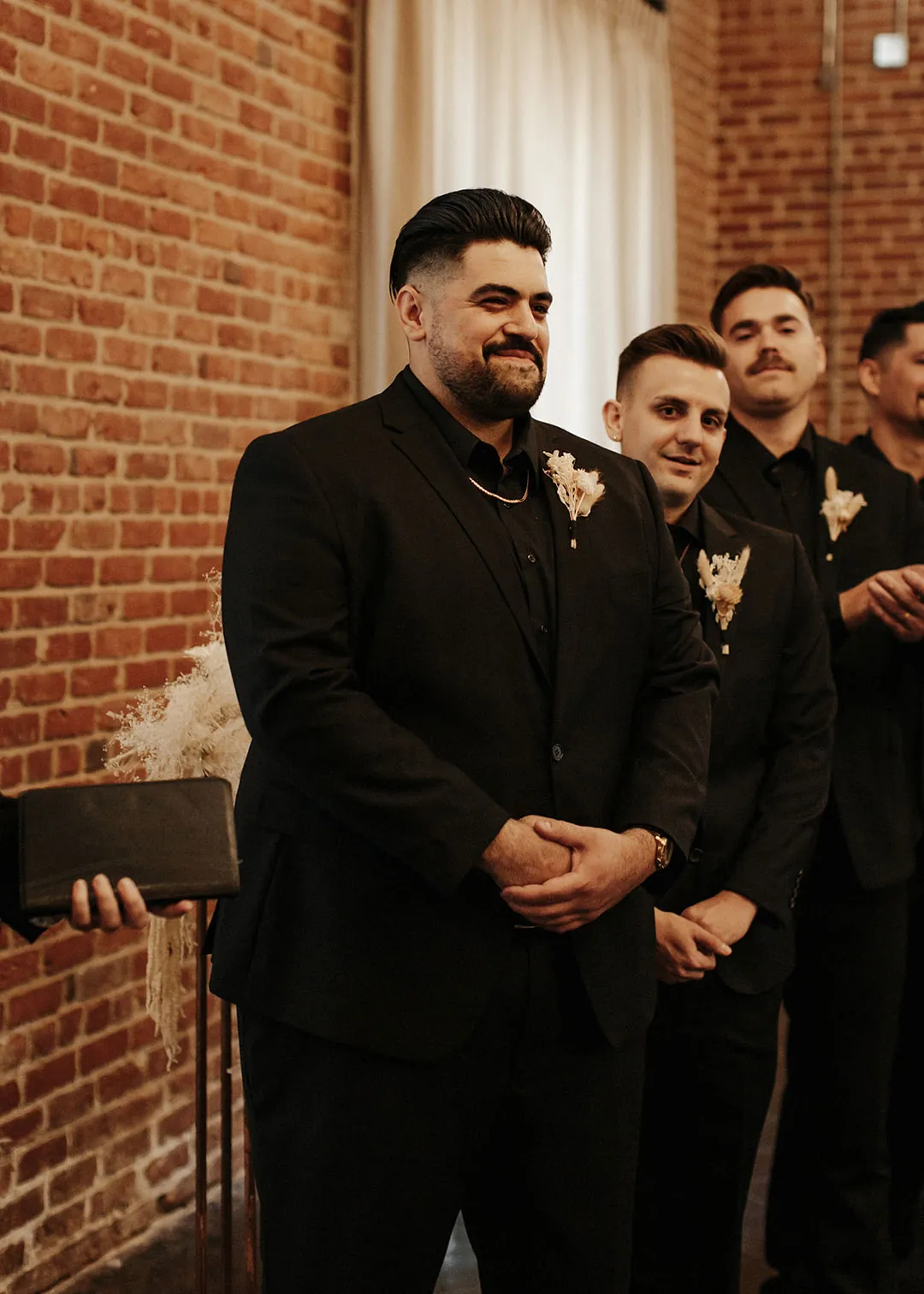 Groom waits with attendants beside exposed brick.
