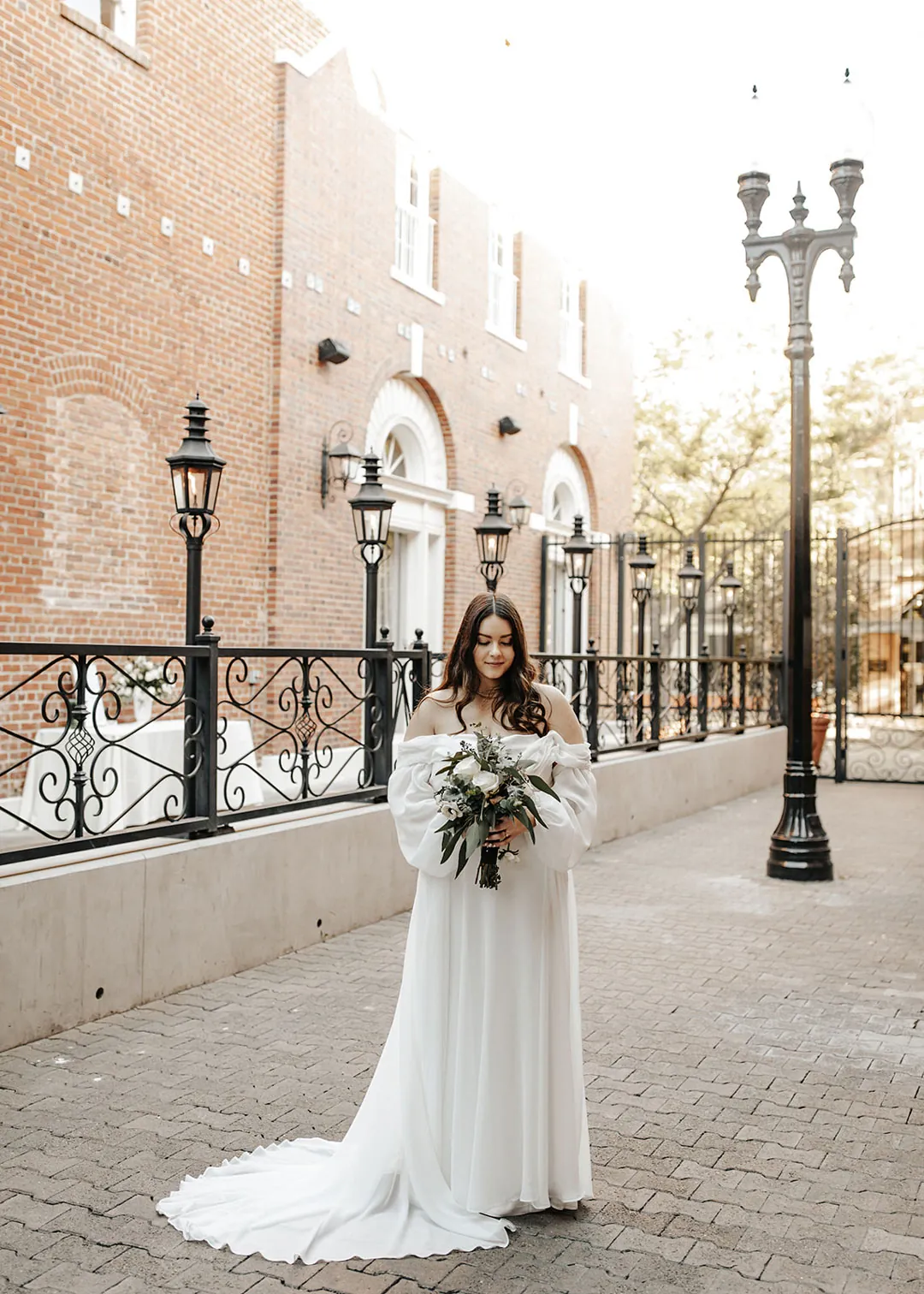 Bride stands along a sunlit brick lined walkway.
