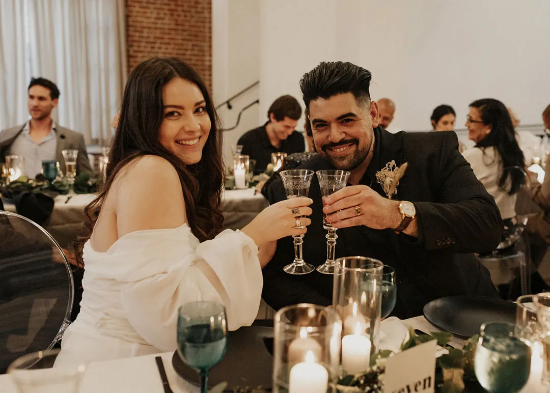 Newlyweds raise glasses while seated at a reception table.