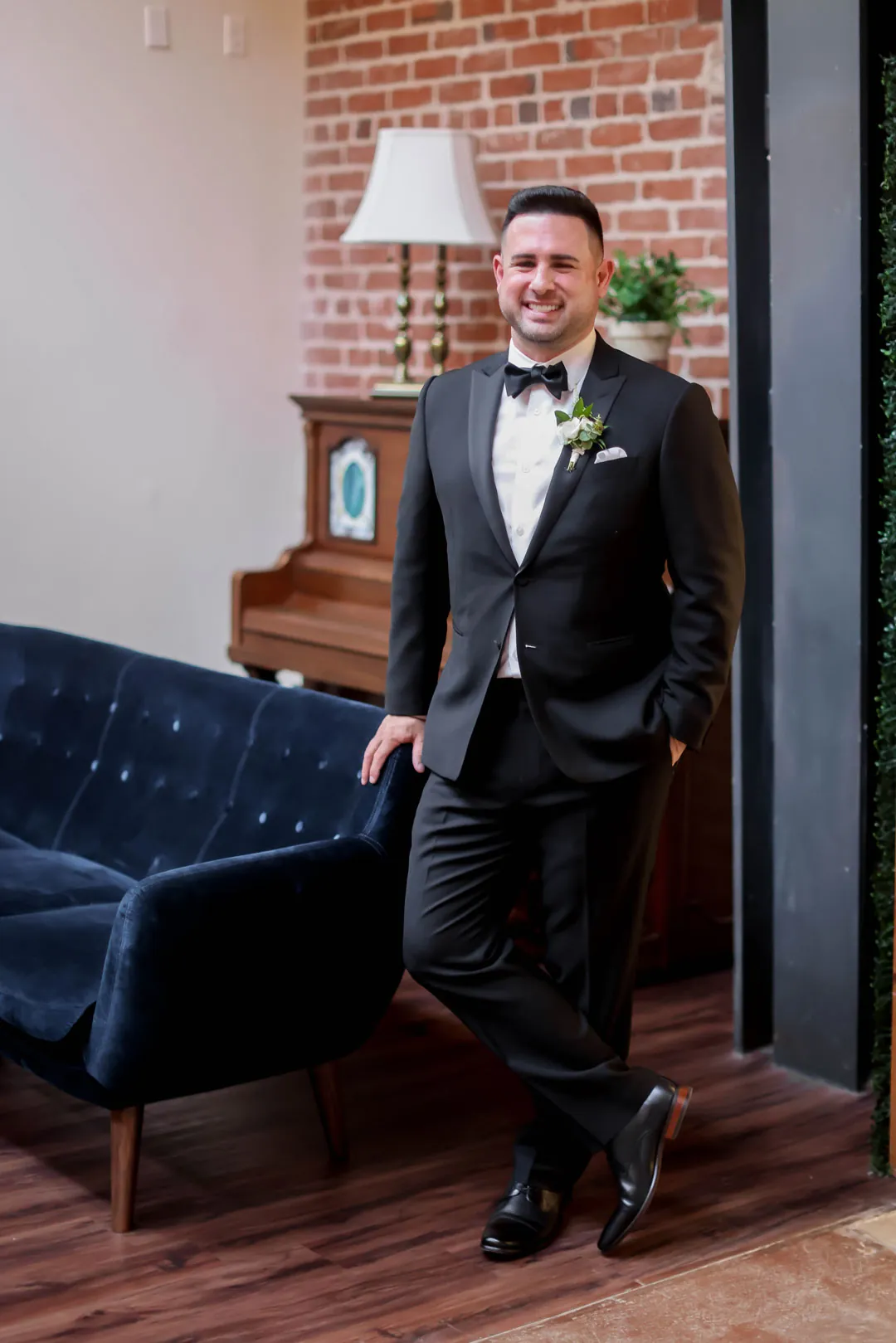 Groom posing in a dark suit beside a sofa