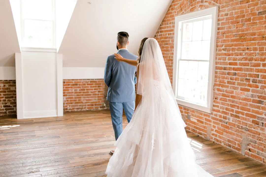 Bride approaching the groom from behind in a sunlit room