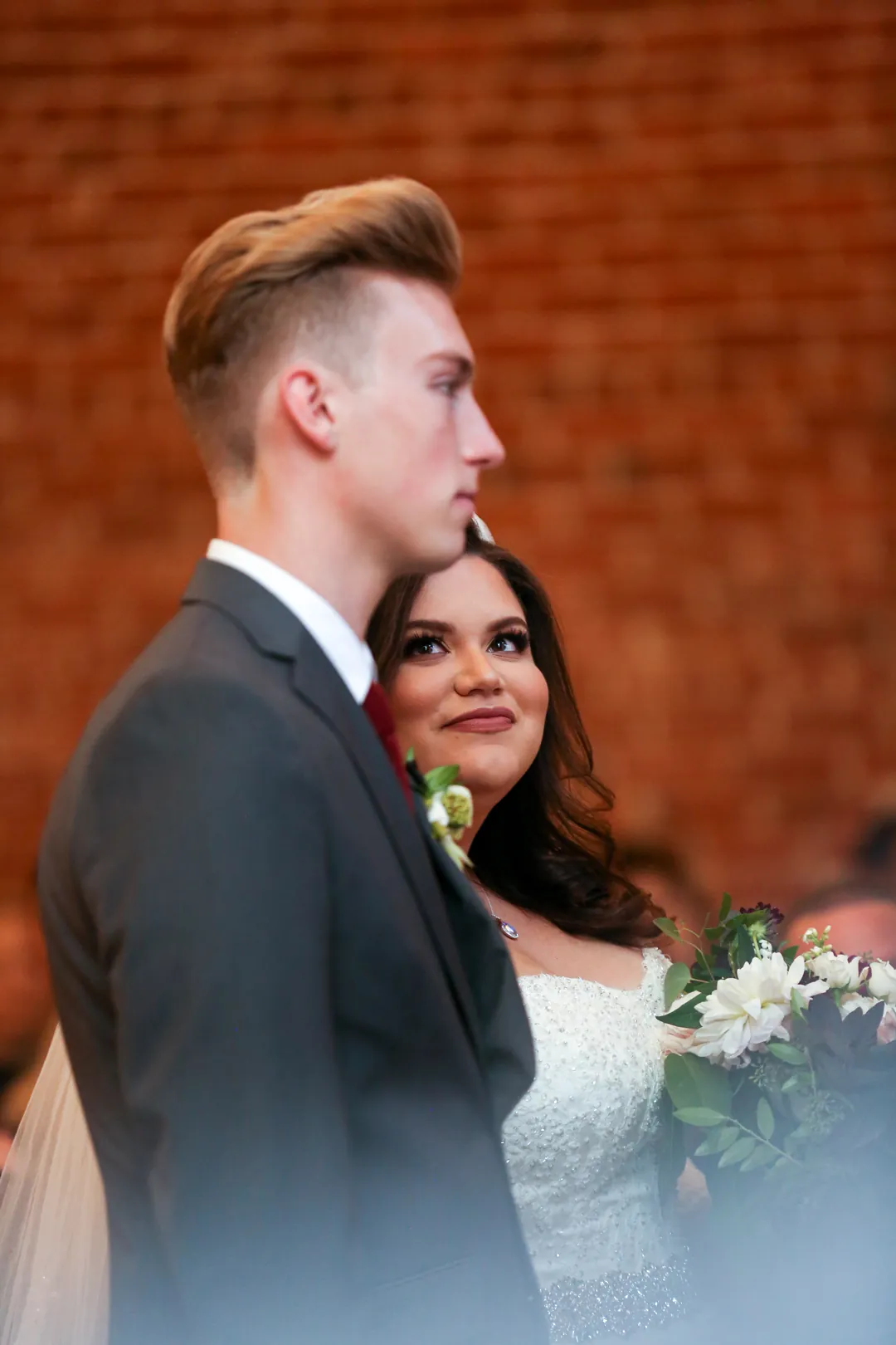 Groom embraces the bride beside exposed brick.
