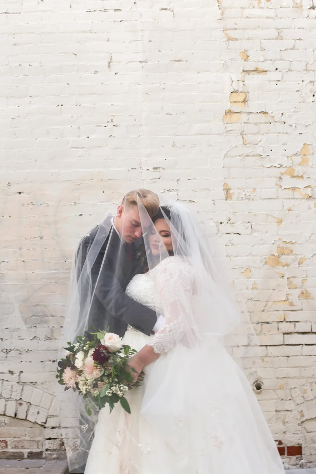 Couple kiss beneath a veil against a white brick wall.