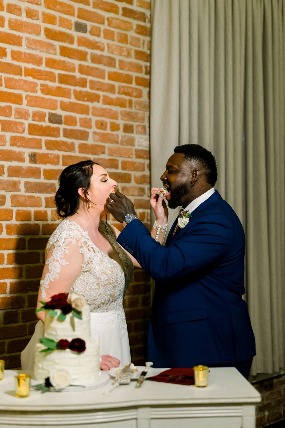 Groom feeding cake to the bride beside a brick wall