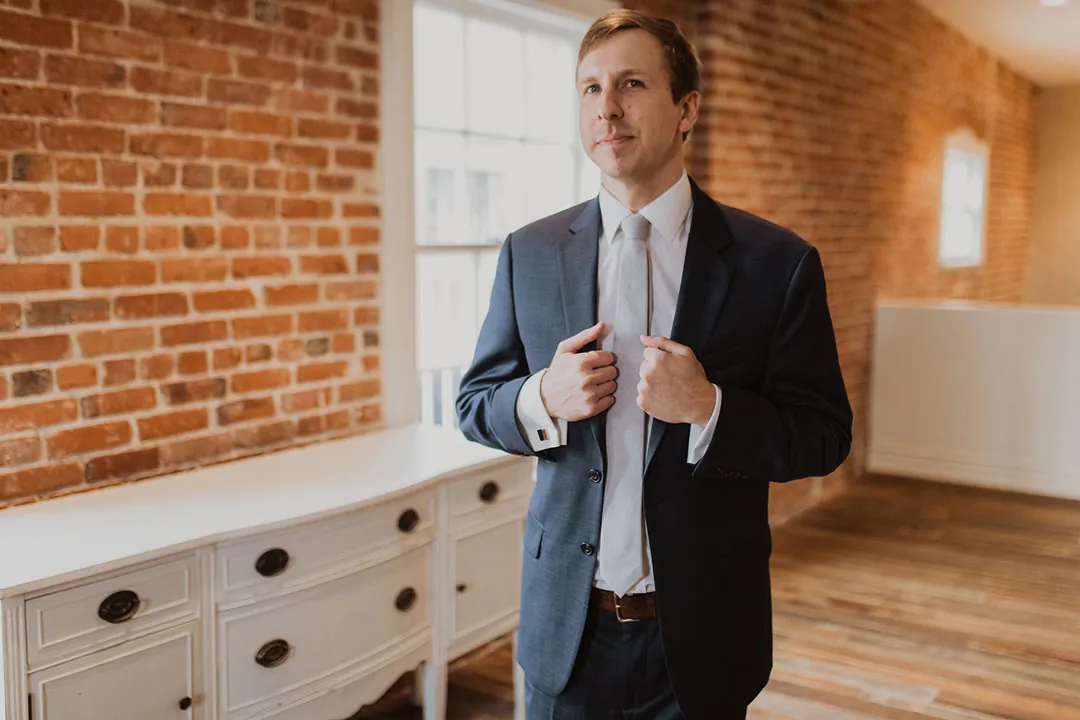 Groom adjusting his jacket beside a loft window
