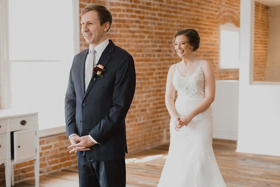 Bride approaching the groom from behind beside a bright window