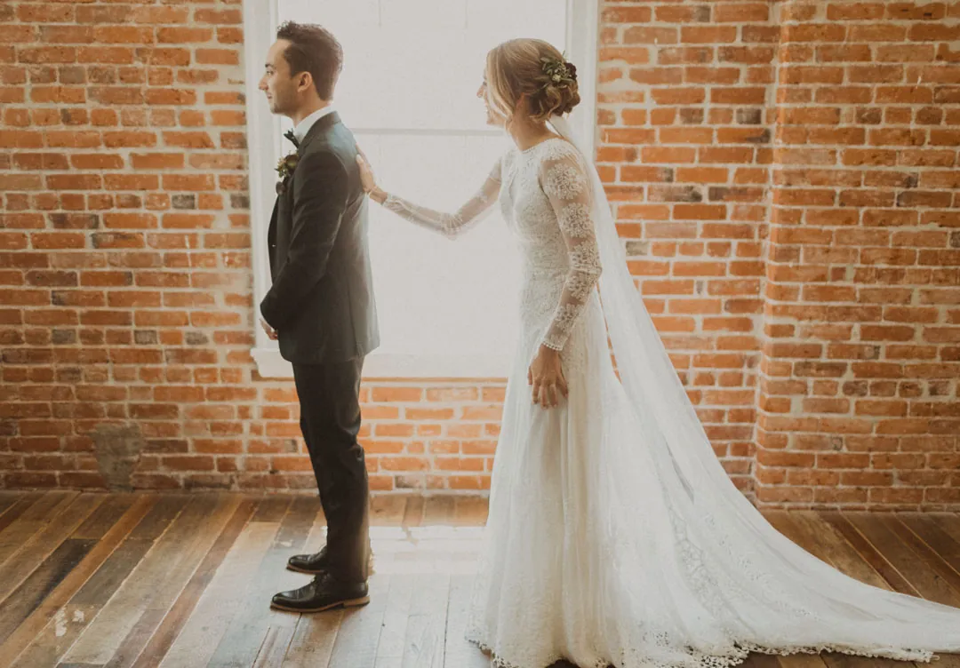 Bride approaching the groom from behind in a brick-walled room
