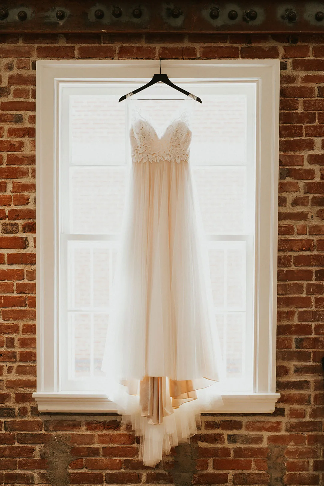 Wedding dress hangs before a tall white window.