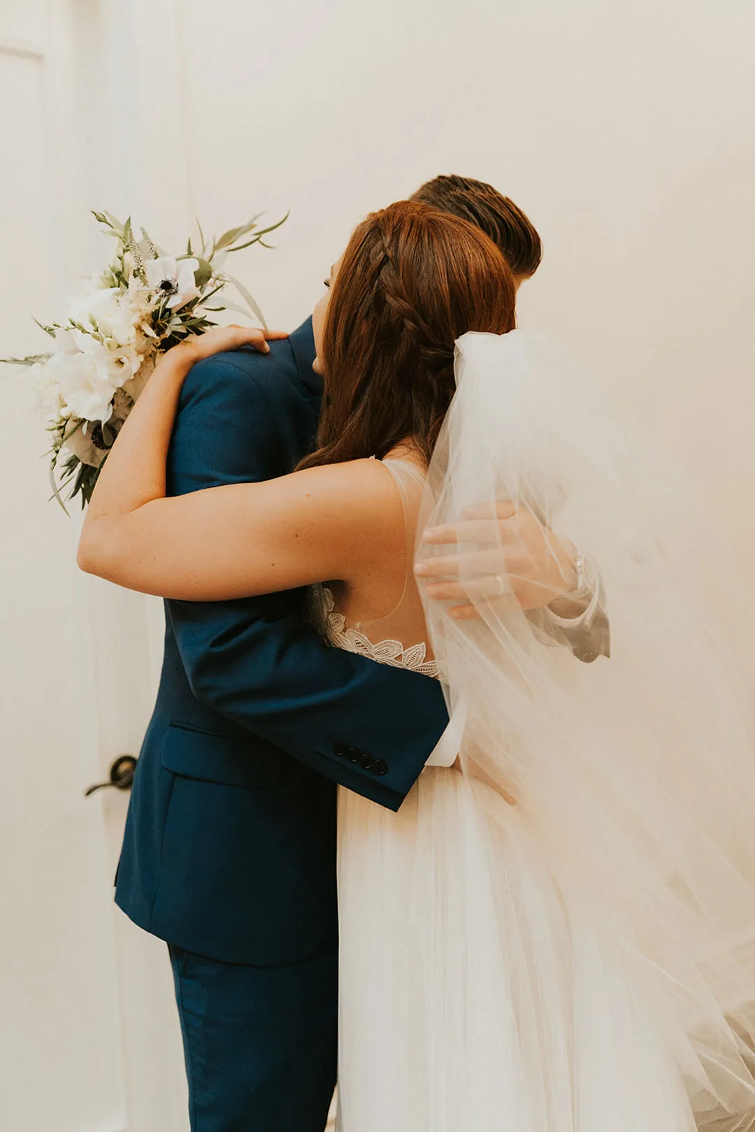 Couple embrace closely against a white wall.