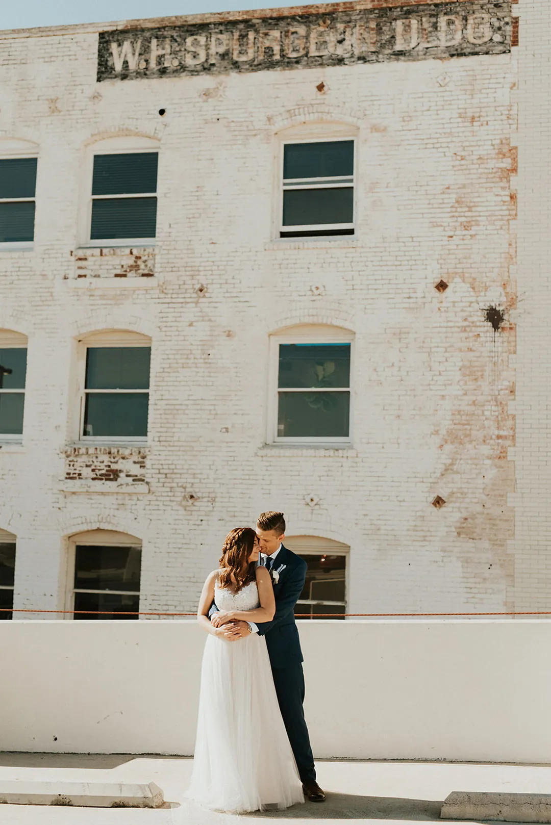 Couple embrace against a pale multistory building.