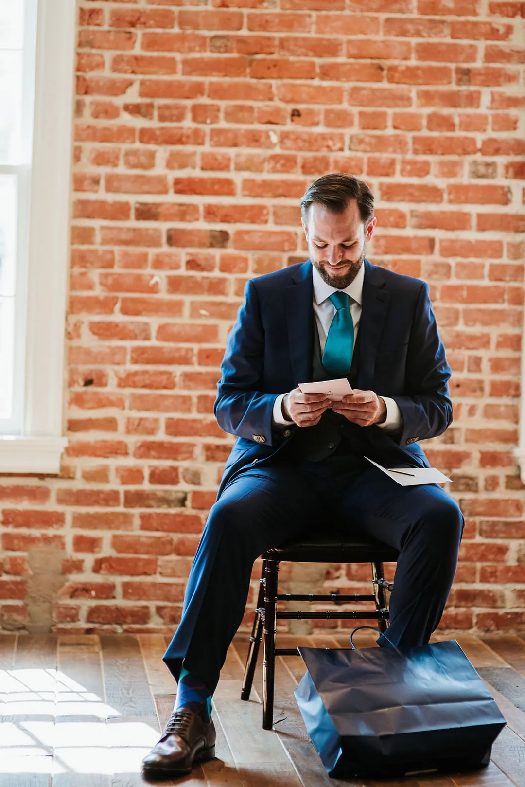 Groom sits beside exposed brick reading a note.